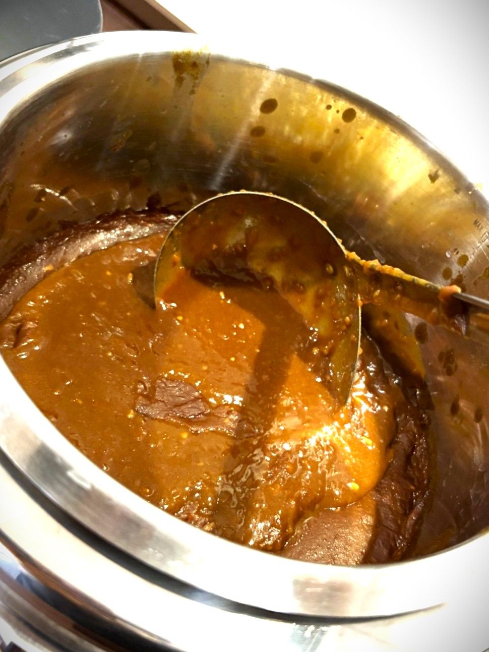 A stainless steel bowl containing a thick, brown sauce with visible chunks, possibly meat, being stirred with a spoon.