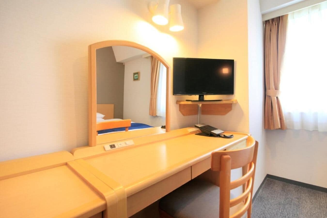 Ein Hotelzimmer mit einem Holztisch, einem Stuhl, einem an der Wand befestigten Fernseher und einem Fenster mit Vorhängen.