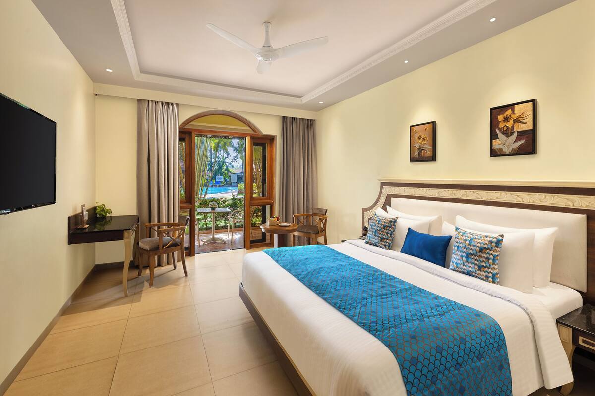 A hotel room with a large bed, a desk with a chair, a TV, and a balcony with a view of palm trees.