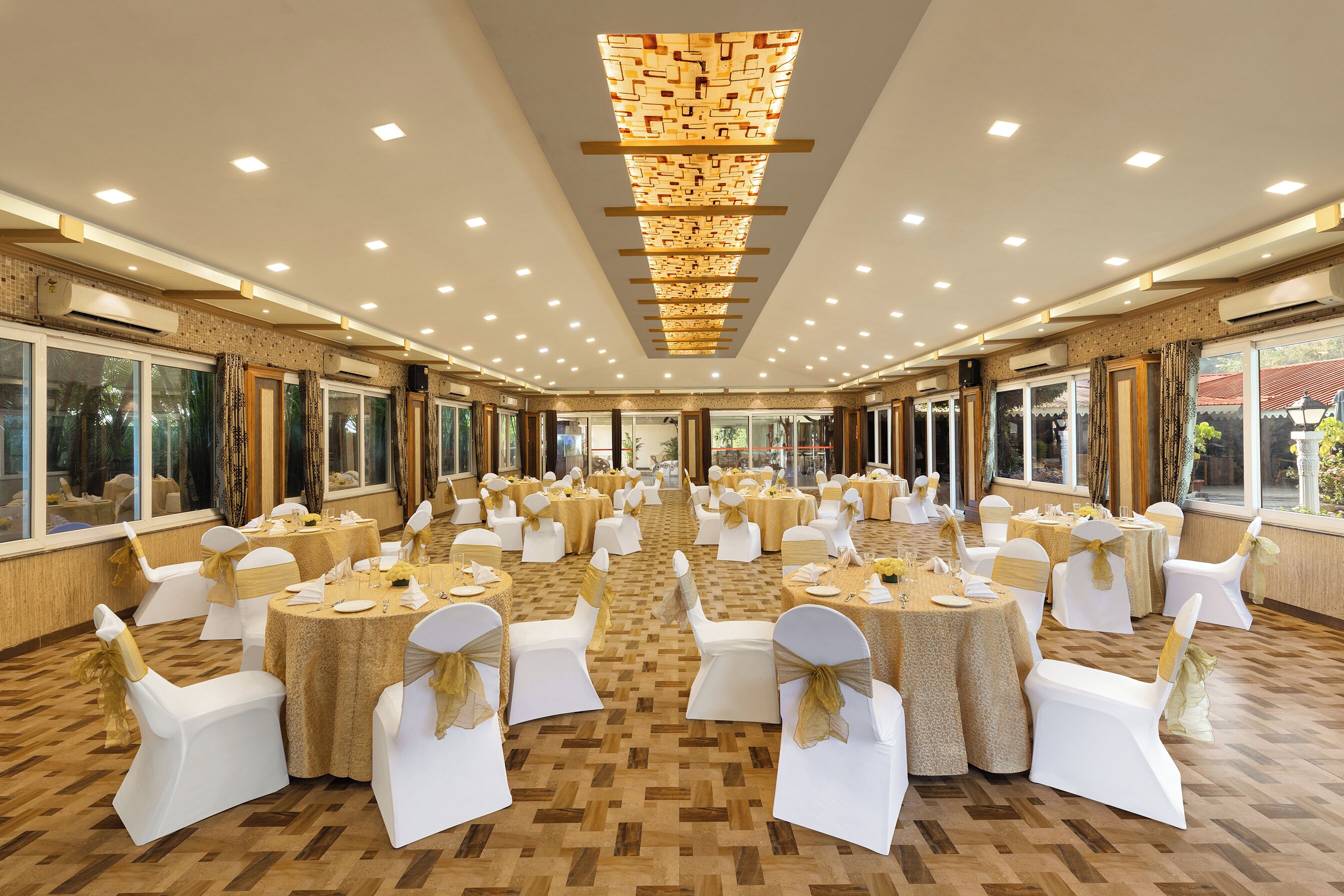 A banquet hall with round tables and white chairs, air conditioning units, and large windows.