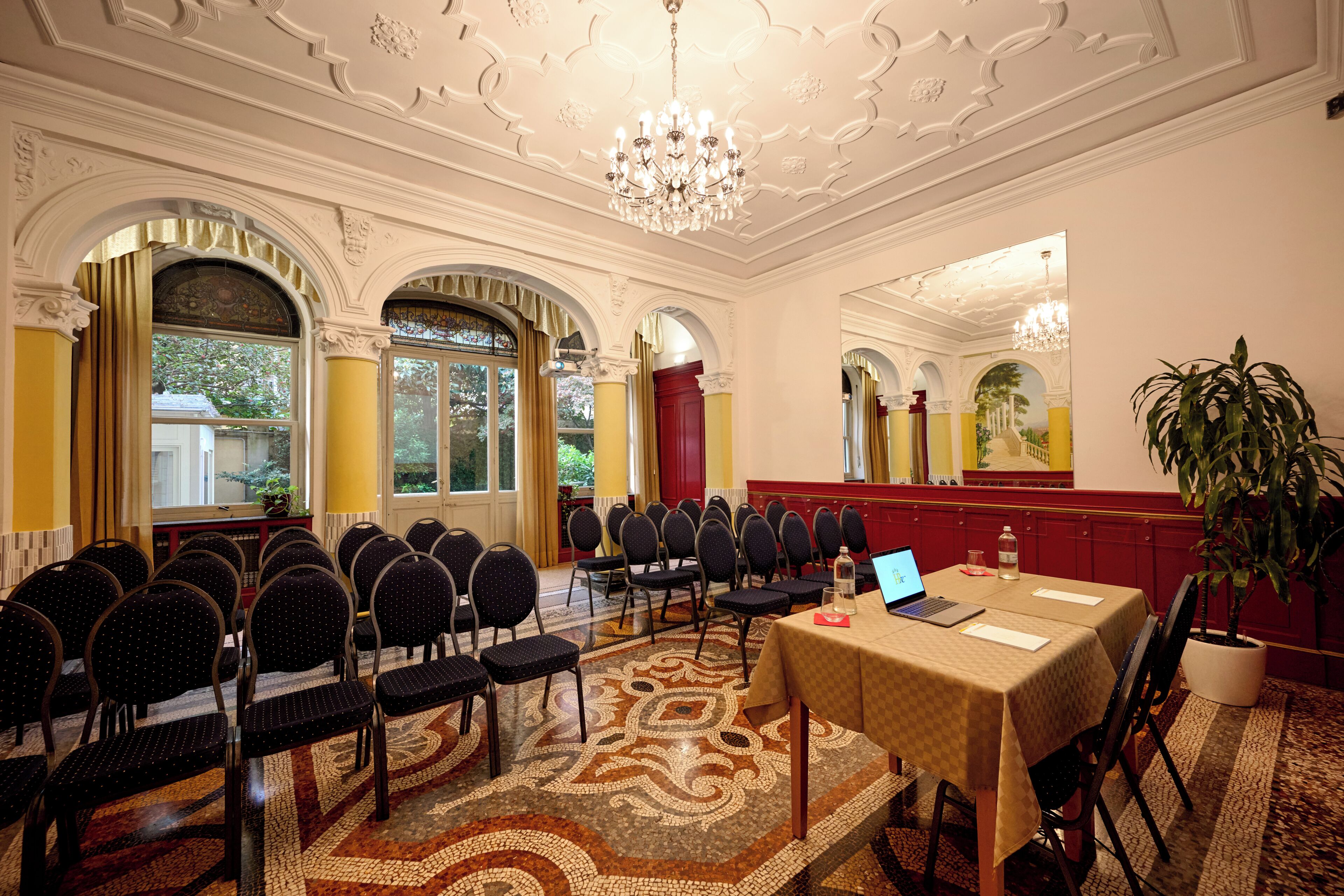A conference room with a laptop on a table, chairs arranged in rows, and a chandelier.