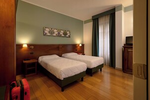 Una camera d'albergo con due letti, una testiera in legno, un comodino, una lampada, un quadro, una televisione e una valigia.