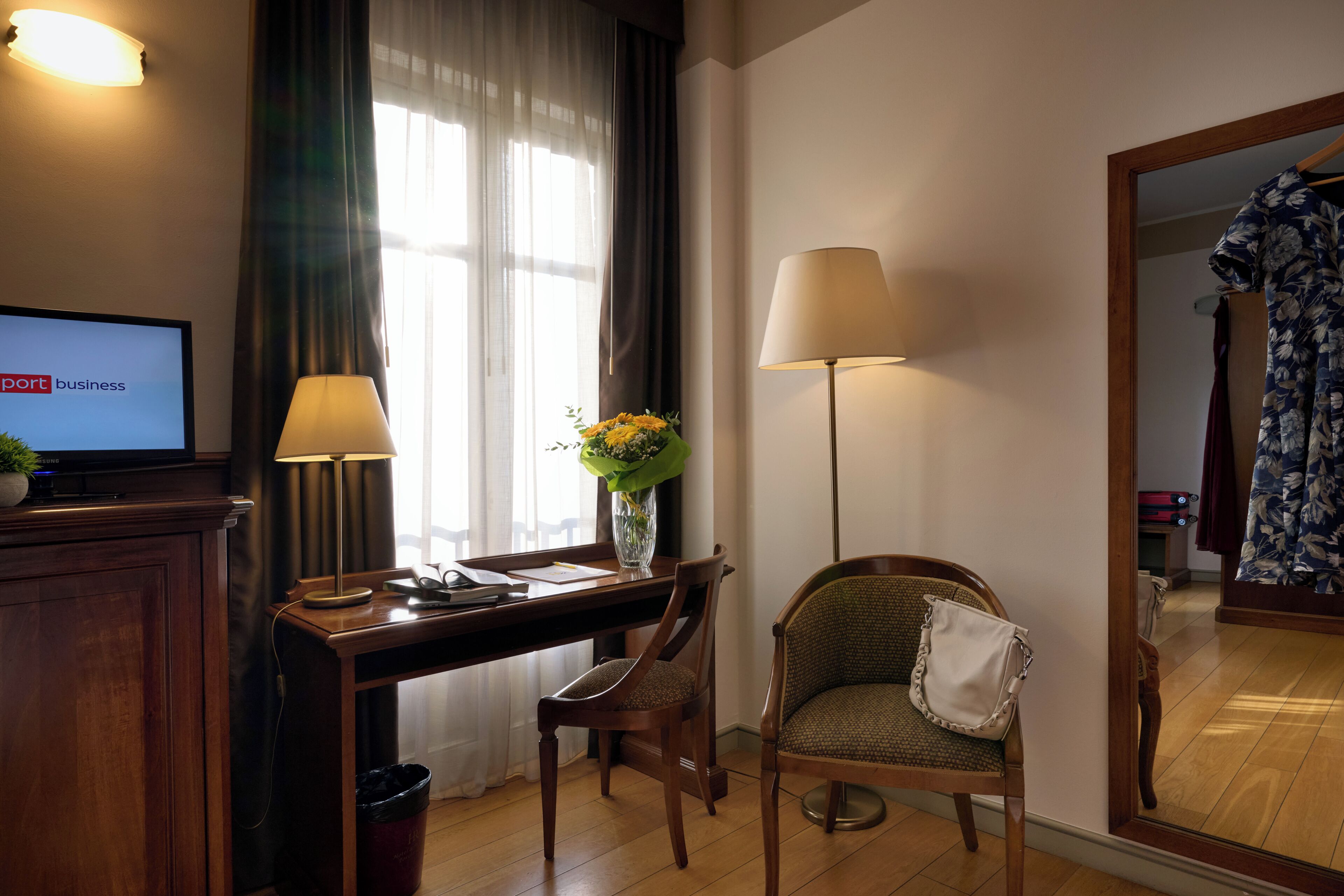 A hotel room with a desk, two lamps, a chair with a bag, a TV displaying 'business', and a mirror reflecting a room with a floral dress.