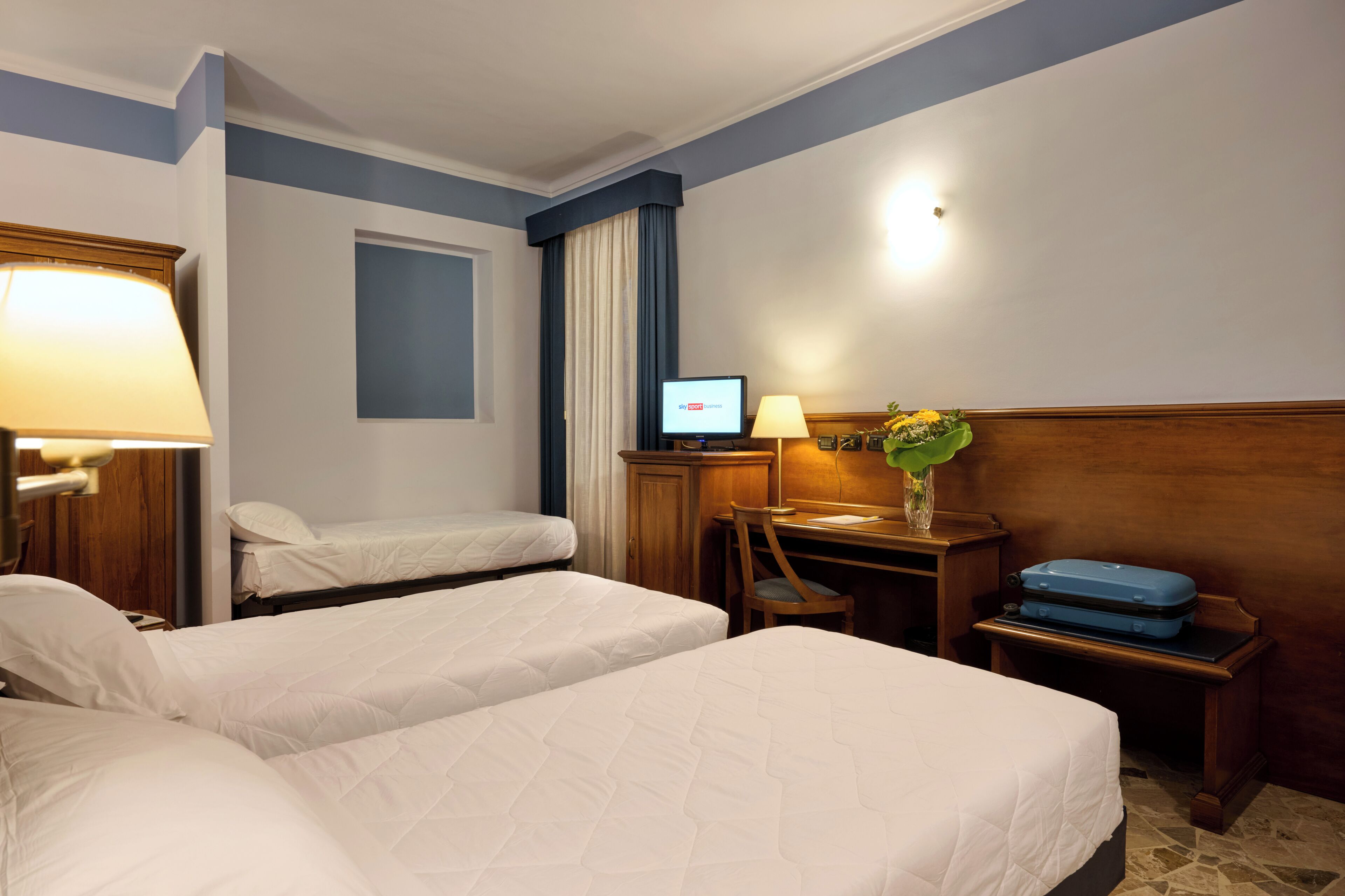 Habitación de hotel con dos camas, un escritorio con ordenador, una lámpara, un florero con flores y una maleta.