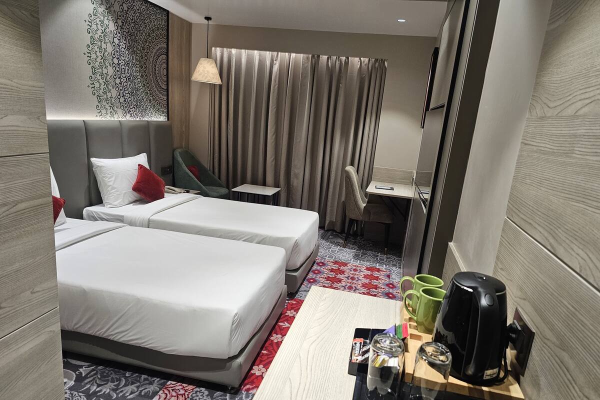 A hotel room with two beds, a desk, a chair, a coffee maker, and a small table with glasses and a bottle.