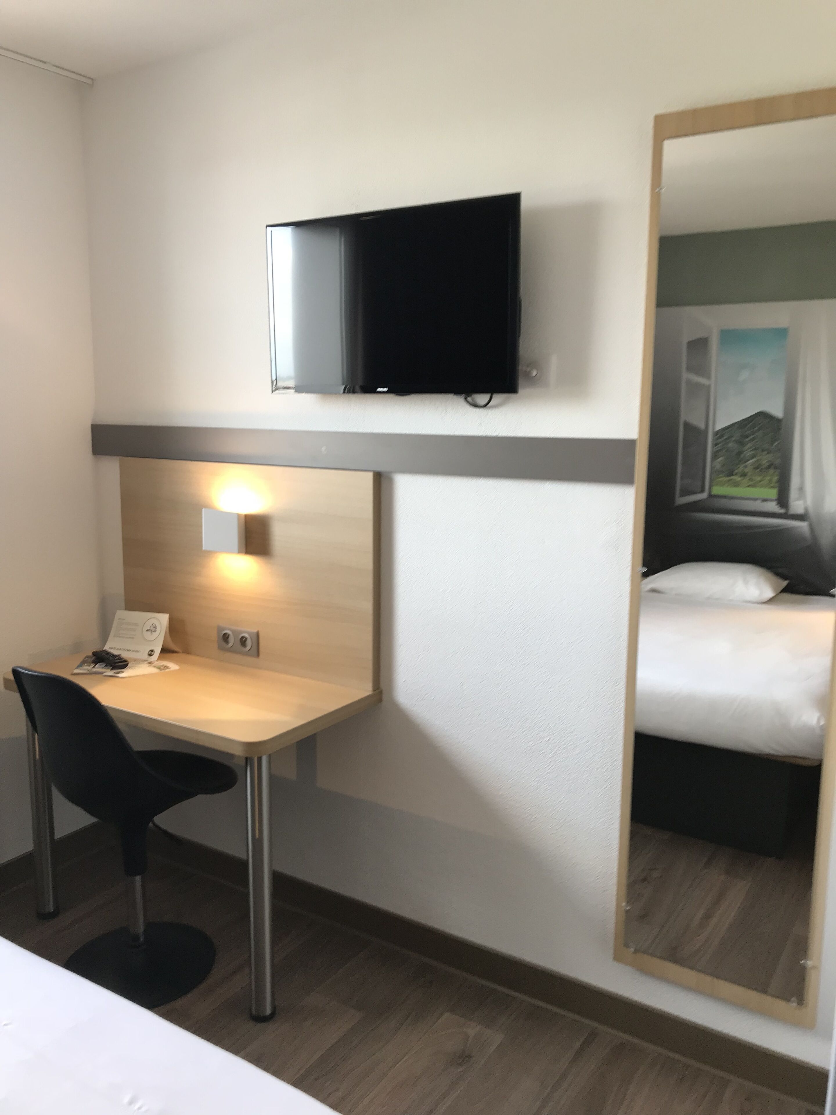 A hotel room with a desk, chair, and a bed. There is a television mounted above the desk and a large mirror reflecting the room.
