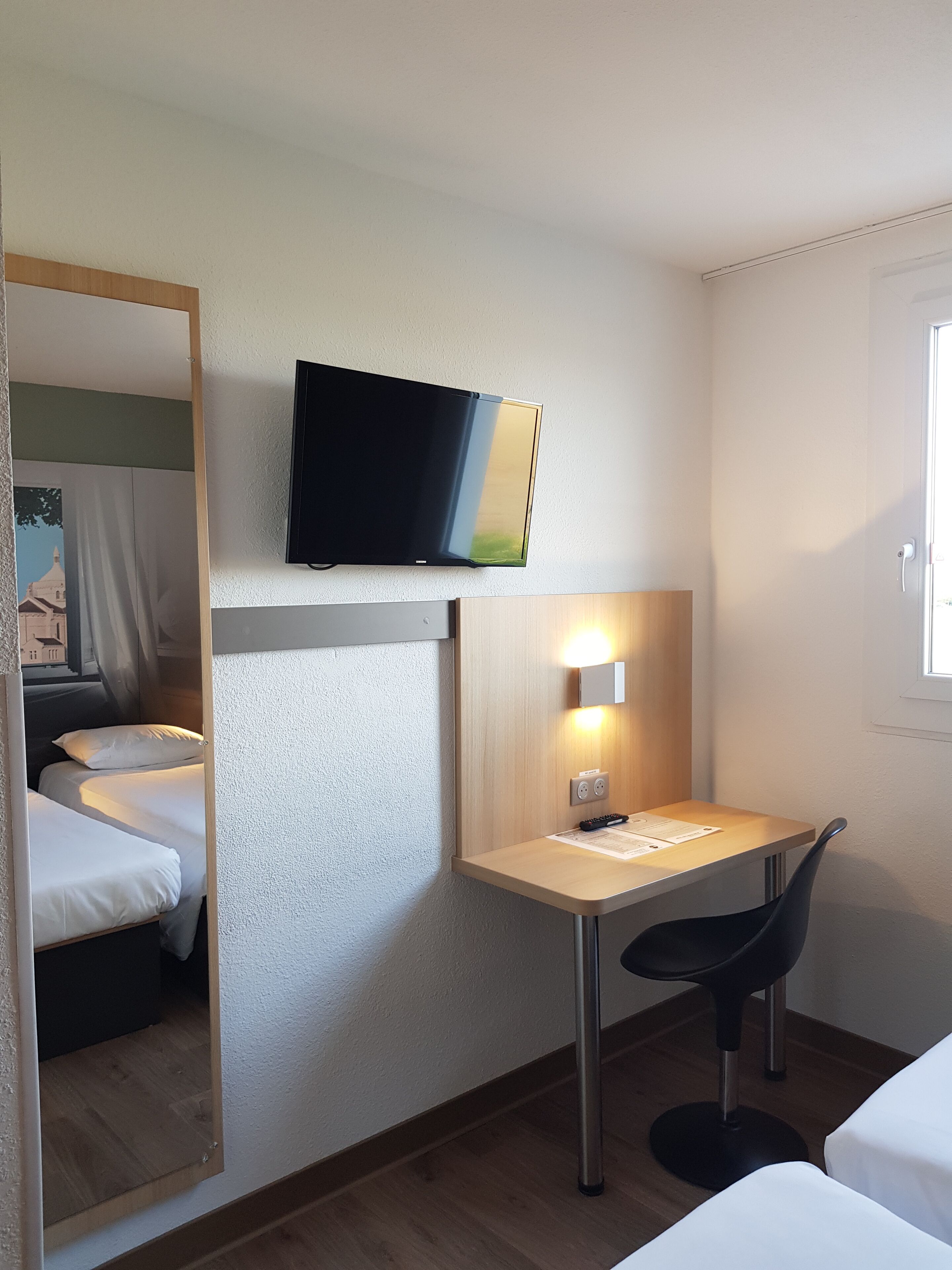 A hotel room with a bed, a desk with a chair, a television mounted on the wall, and a window.
