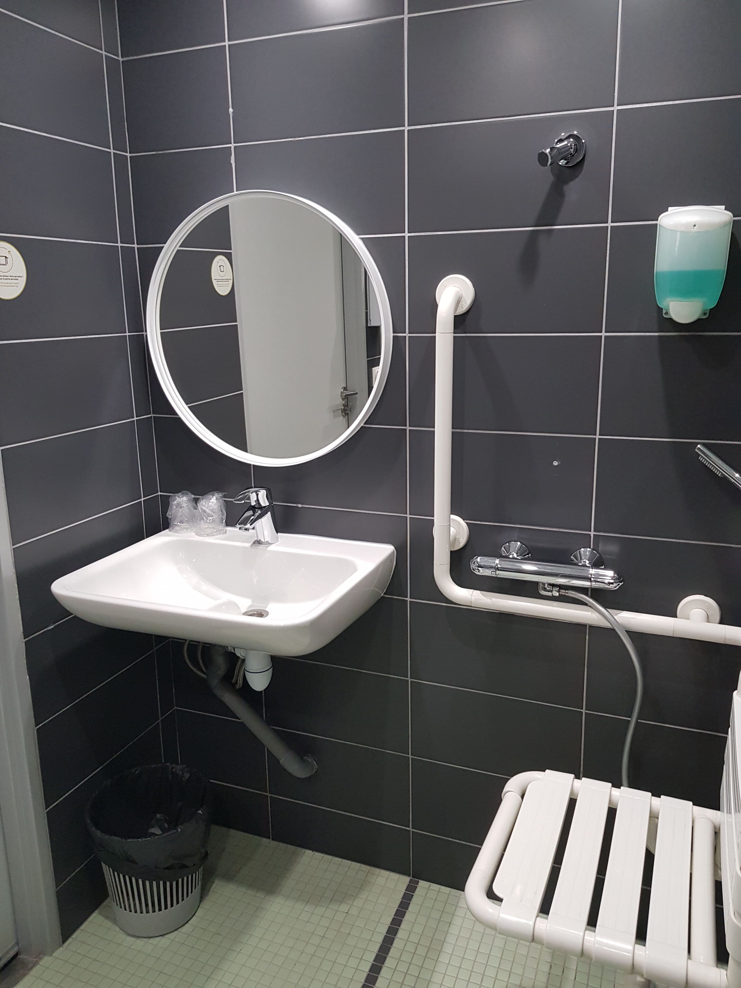 Ein Badezimmer mit einem weißen Waschbecken, einem Spiegel, einem Händetrockner und einem Papierkorb.