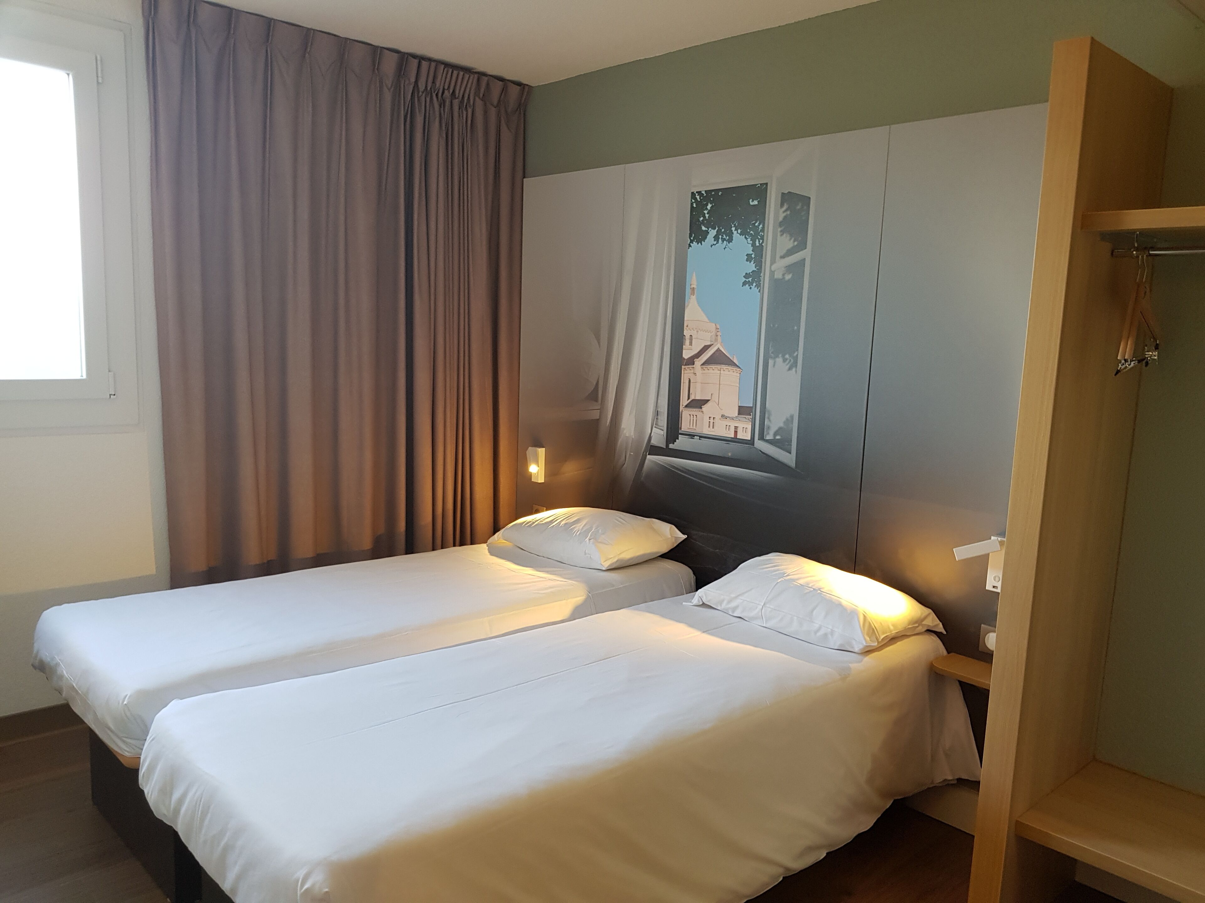 Ein Hotelzimmer mit zwei Einzelbetten, einem großen Fenster mit Vorhängen, einer Wandlampe und einem Holzregal.