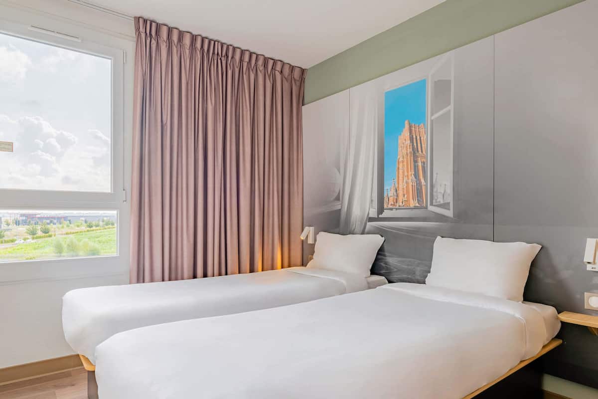 Une chambre d’hôtel avec deux lits, une grande fenêtre avec des rideaux, un tableau représentant un bâtiment et une vue sur la verdure à l’extérieur.