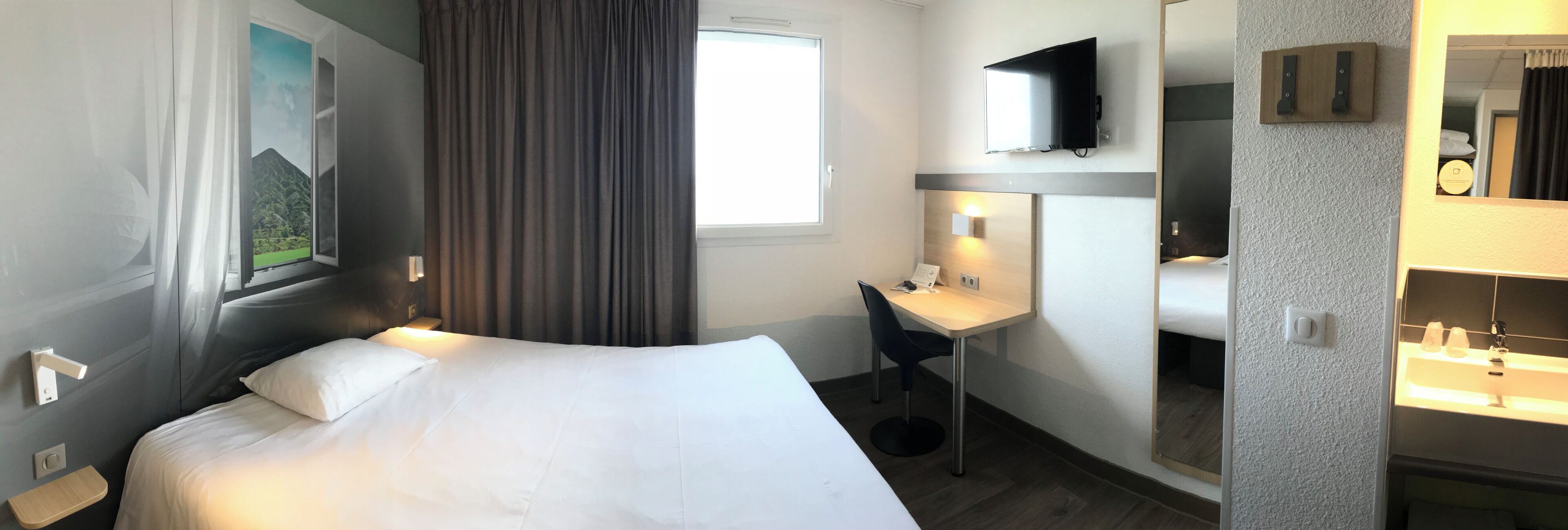 A hotel room with a bed, a desk, a TV, a mirror, and a bathroom.