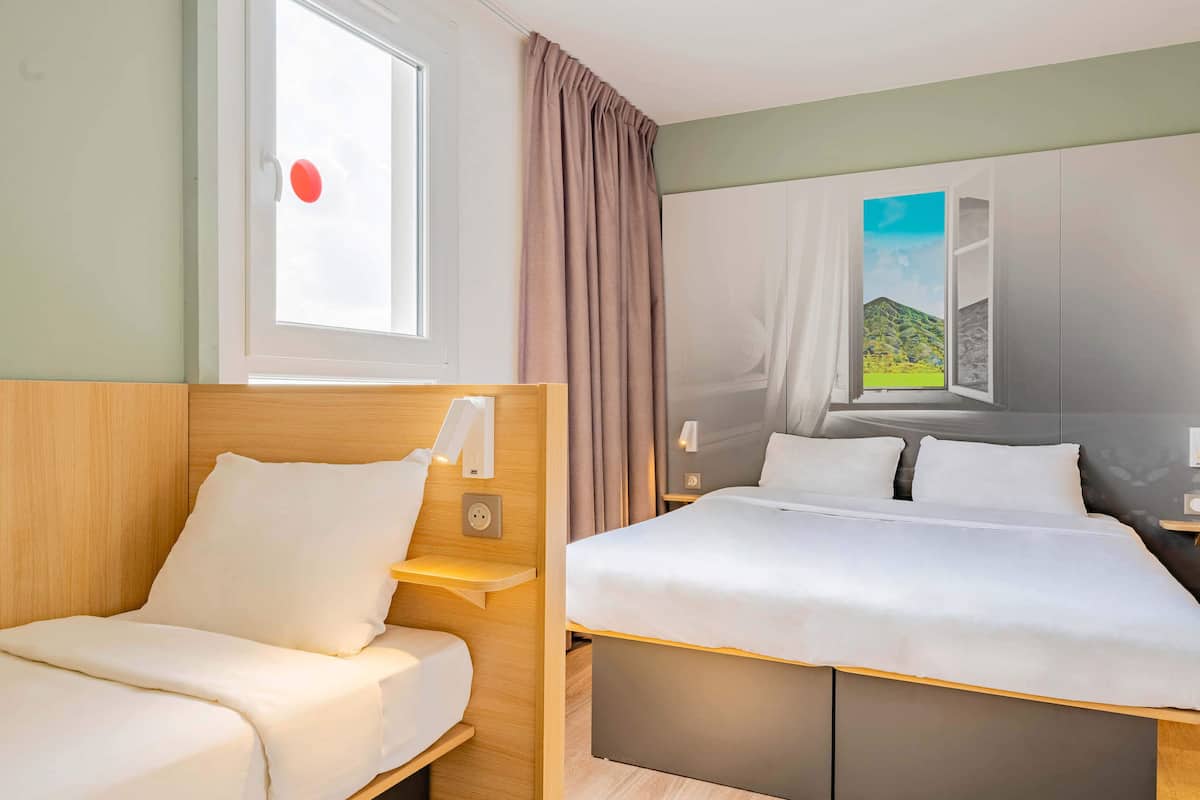 Une chambre d’hôtel avec deux lits, une fenêtre avec des rideaux, une lampe fixée au mur et un tableau représentant un paysage.