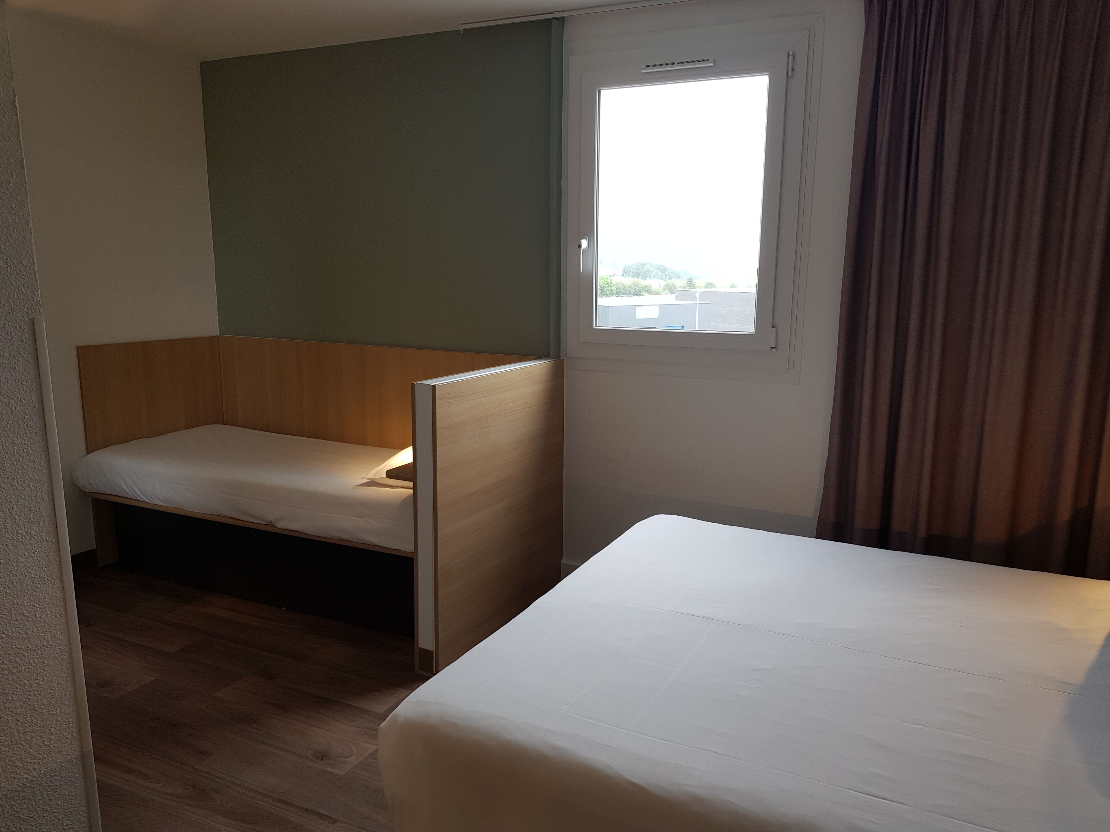 Ein Hotelzimmer mit einem Einzelbett, einer kleinen Holzbank, einem Fenster mit Vorhängen und Blick nach draußen.