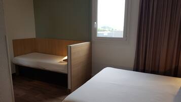 Habitación de hotel con una cama individual, un pequeño banco de madera, una ventana con cortinas y vistas al exterior.