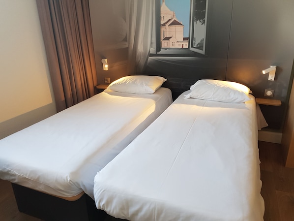 A hotel room with two single beds, a nightstand with a lamp, and a view of a building through a window.