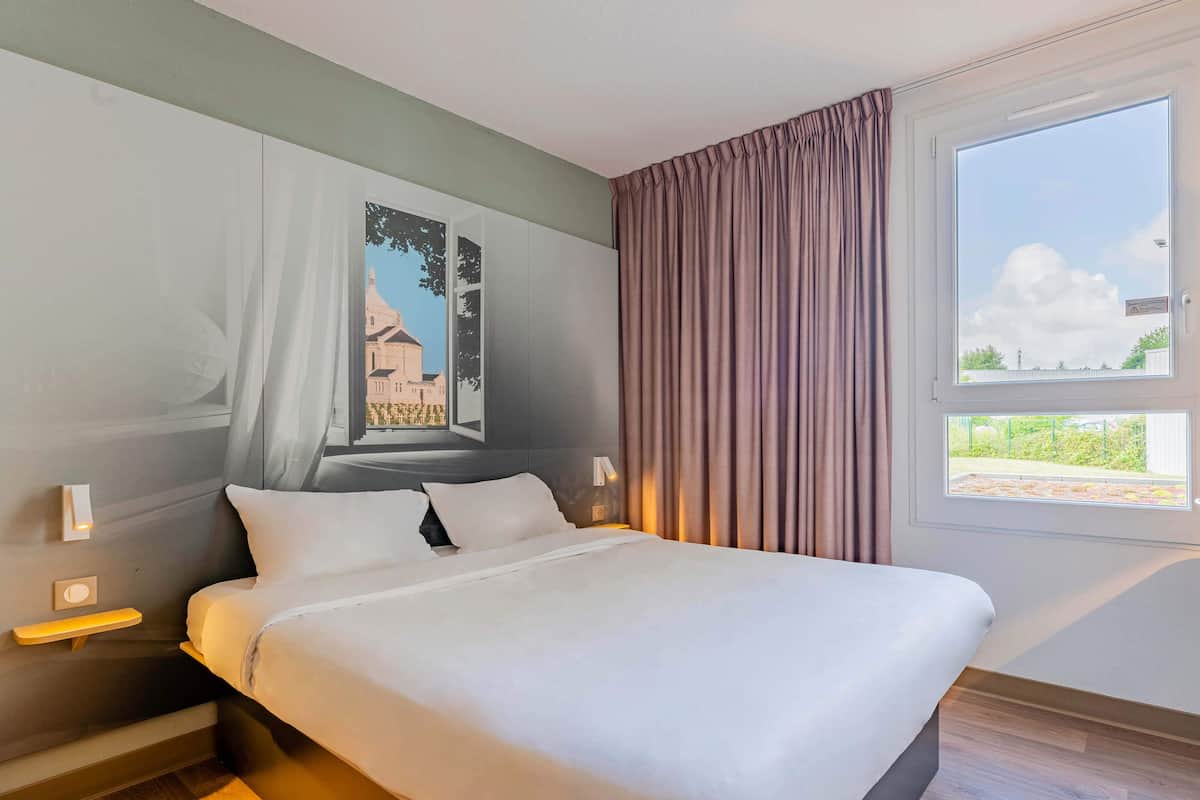 Un lit bien fait dans une chambre d’hôtel, avec une grande fenêtre, une table de chevet et une lampe fixée au mur.