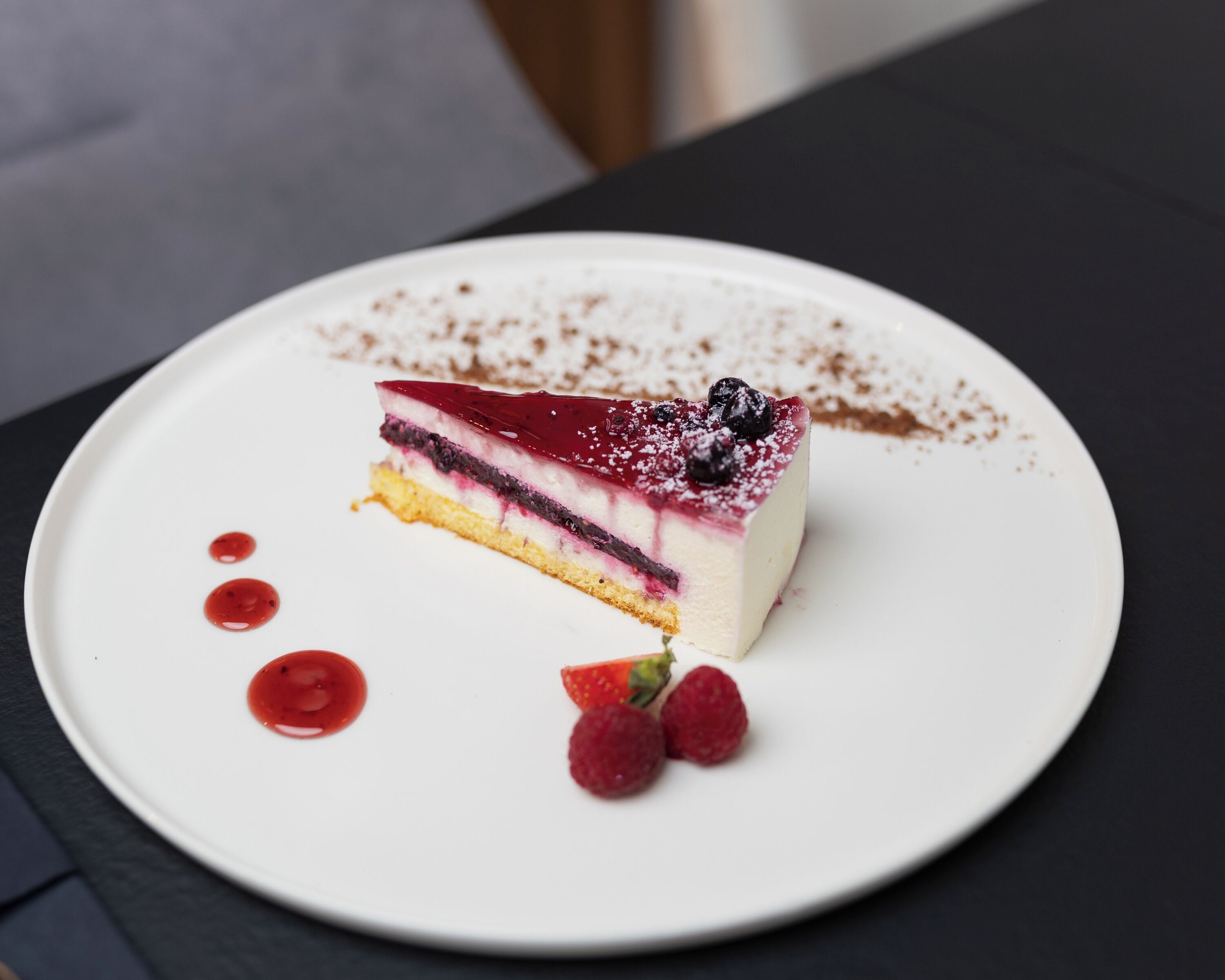Una fetta di dolce stratificato su un piatto bianco, guarnita con frutti di bosco e una spolverata di zucchero, posata su una superficie scura.