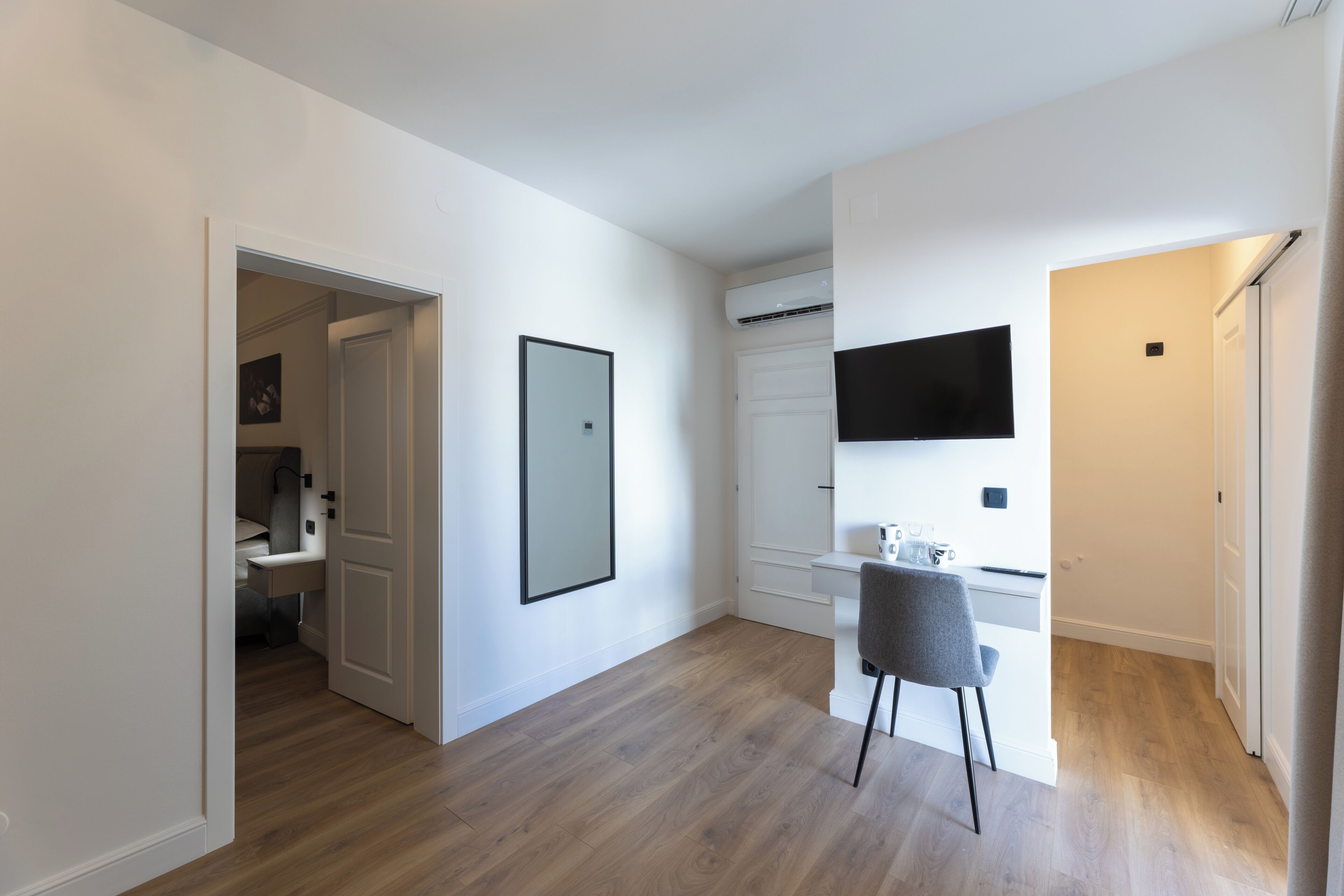 A modern, well-lit room with a wooden floor, a desk with a chair, a mounted TV, and a door leading to another room.