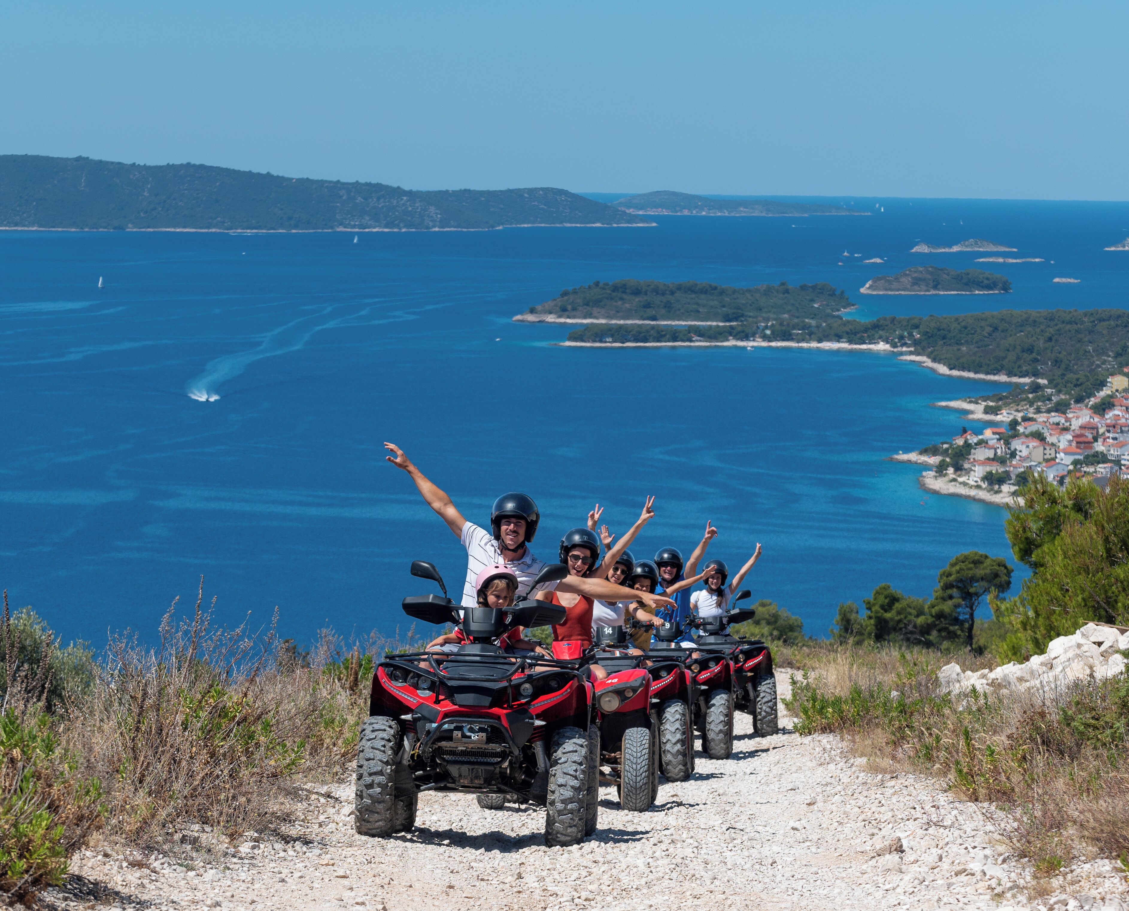 Egy csoport ember, akik négykerekű motoros járműveken (ATV-ken) haladnak egy poros úton, ahonnan a tengerpartra nyílik kilátás.
