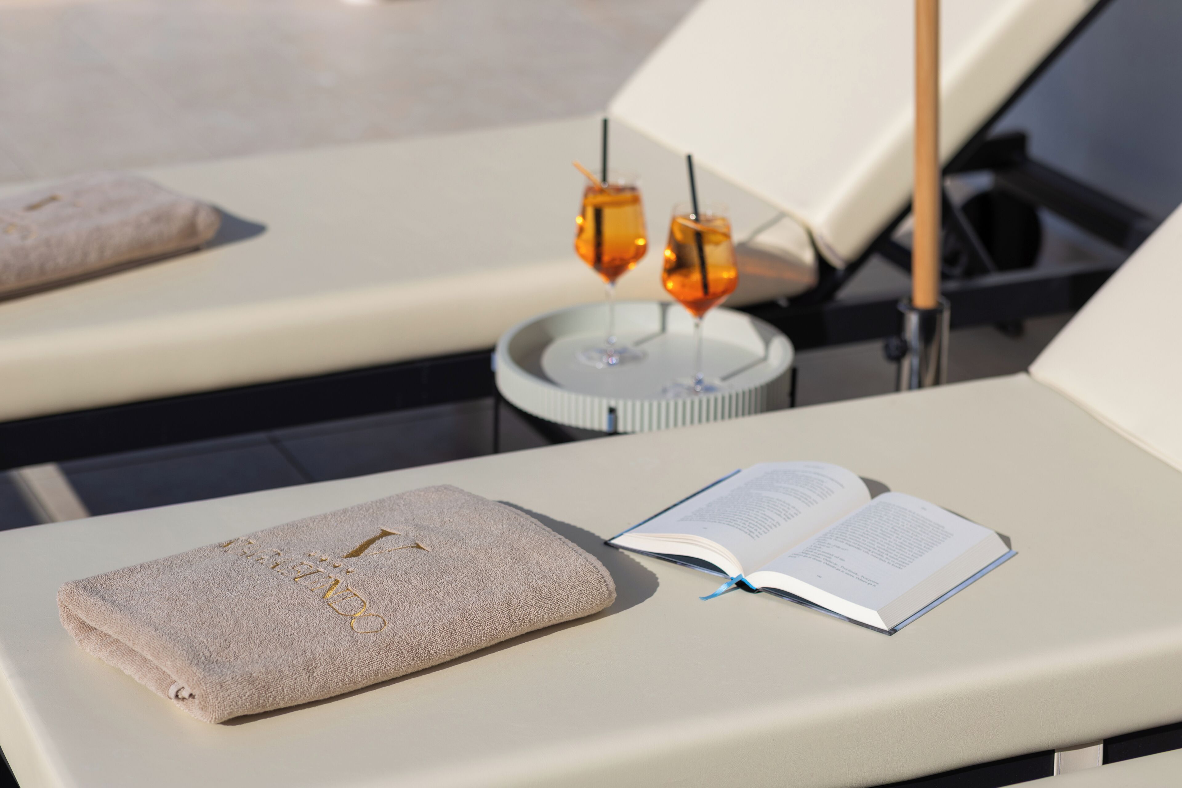 A sun lounger with a towel and an open book, a small table with two drinks, and a poolside view.