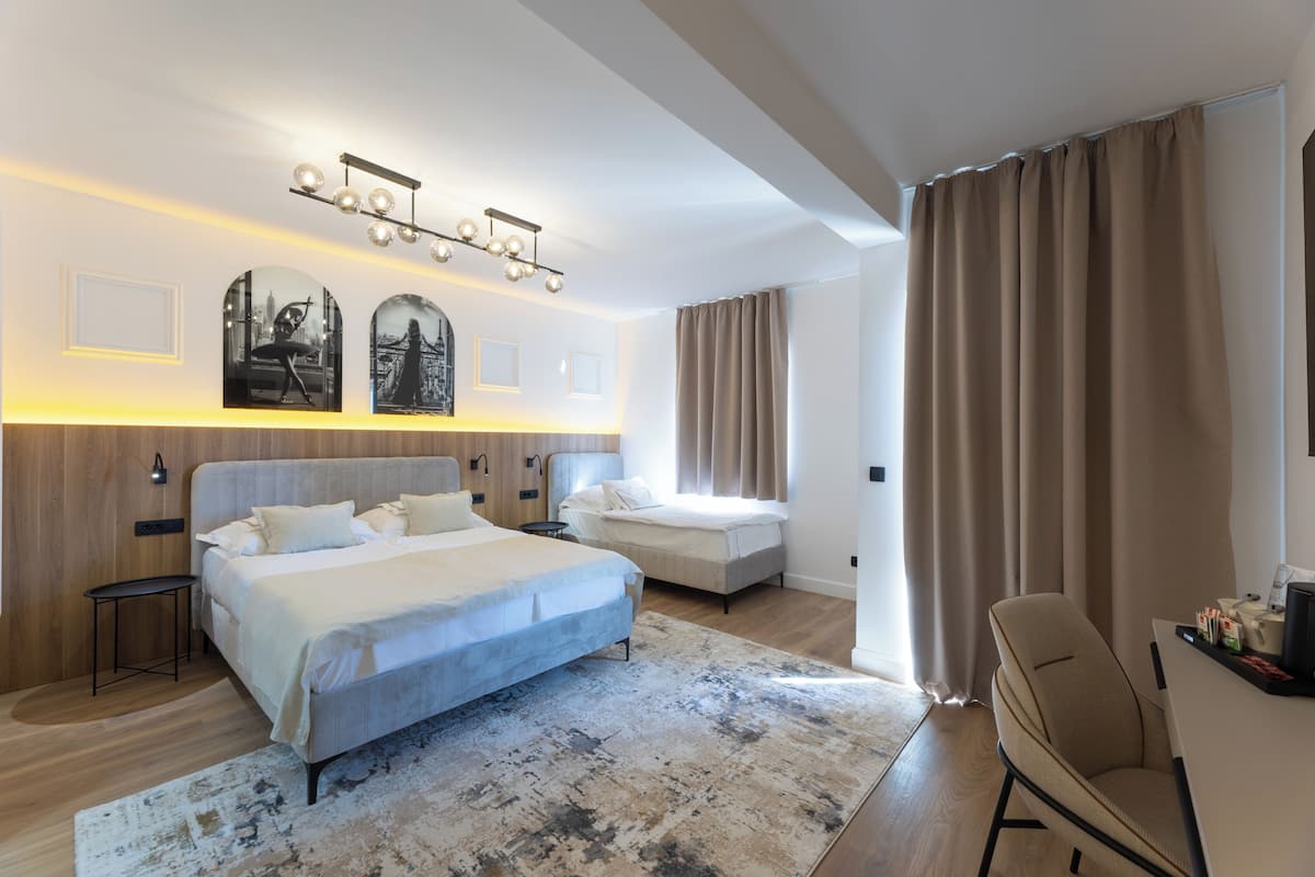 Quarto de hotel moderno com uma cama grande, uma escrivaninha e uma cadeira. Há duas luminárias fixadas na parede e uma janela ampla com cortinas.