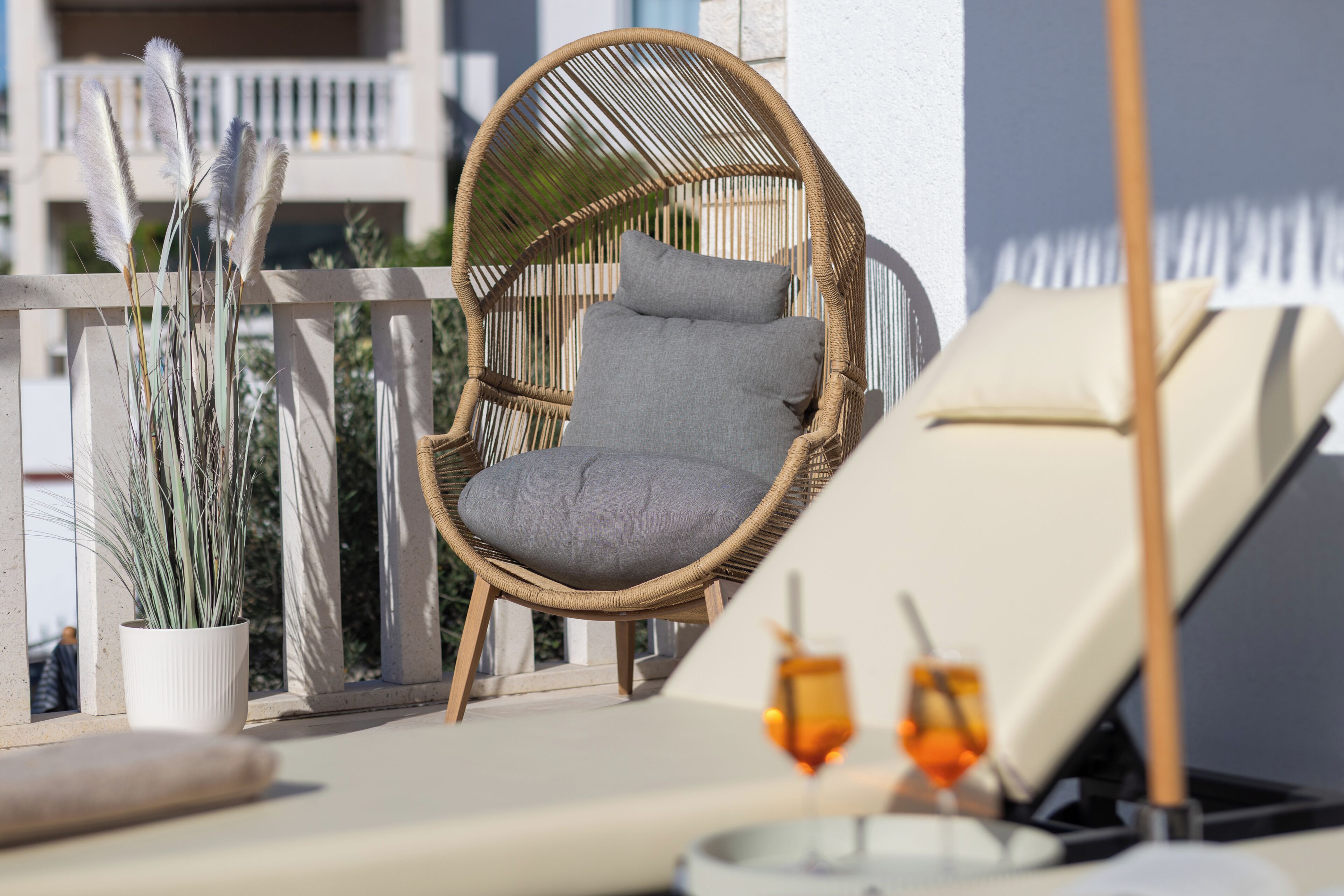 A wicker chair with cushions, a lounge chair, a potted plant, and two glasses of drink on a balcony.