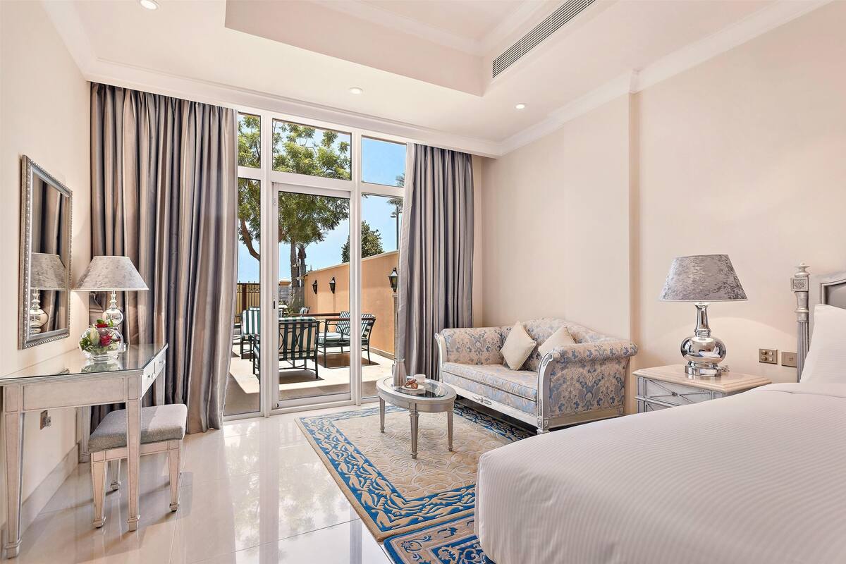 A hotel room with a bed, a sofa, a desk with a lamp, a mirror, and a view of the outside.