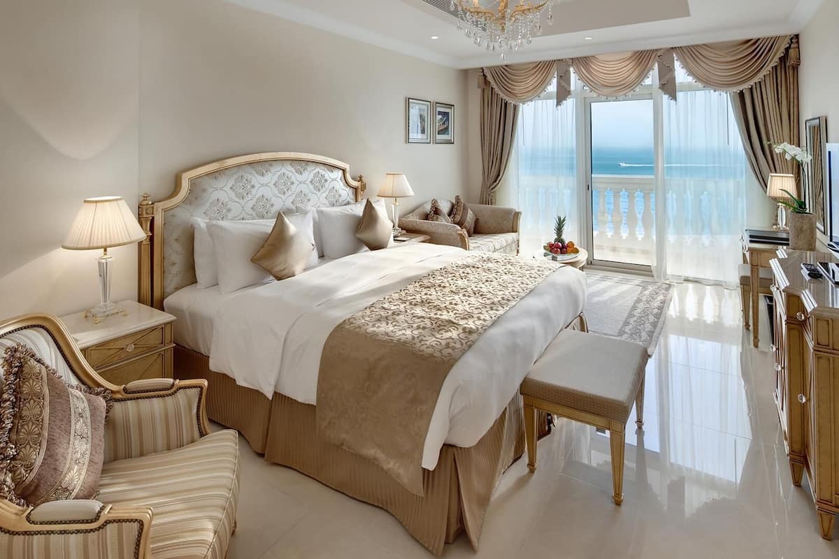 A luxurious bedroom with a large bed, a sitting area with a chaise lounge, a television, and a view of the sea.