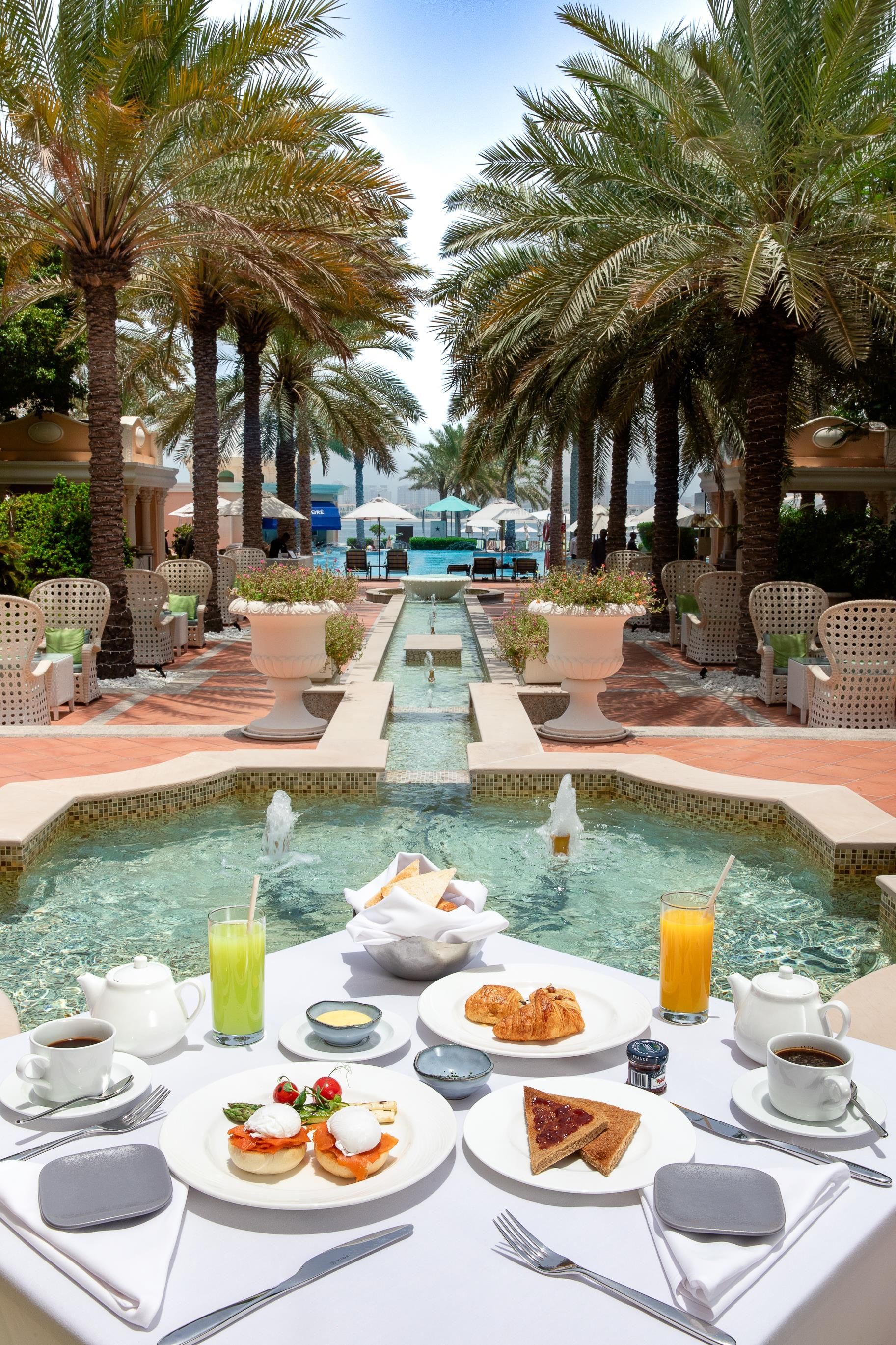 A poolside dining area with a table set for breakfast, surrounded by palm trees and a view of a swimming pool.