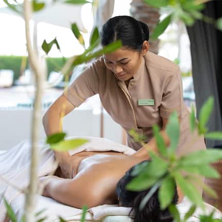 A massage therapy session in a spa setting with a client lying on a massage table, a therapist attending to the client, and a view of the outdoor pool area in the background.