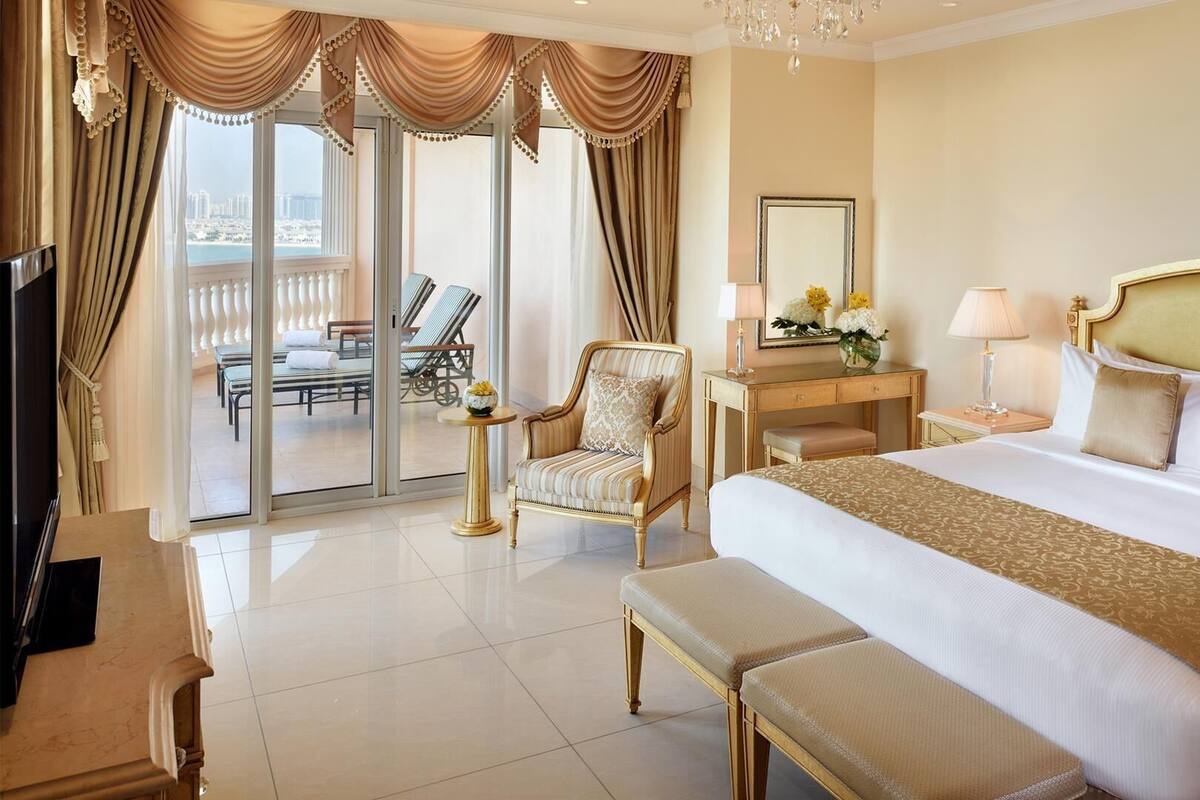 A spacious hotel room with a large bed, a sitting area with a chair and a table, a television, and a balcony with outdoor seating.