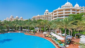 A large swimming pool with lounge chairs and umbrellas, surrounded by palm trees and a grand hotel building with multiple domes and balconies.