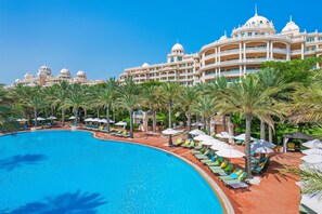 Una gran piscina con sillas de descanso y sombrillas, rodeada de palmeras y un imponente edificio de hotel con múltiples cúpulas y balcones.