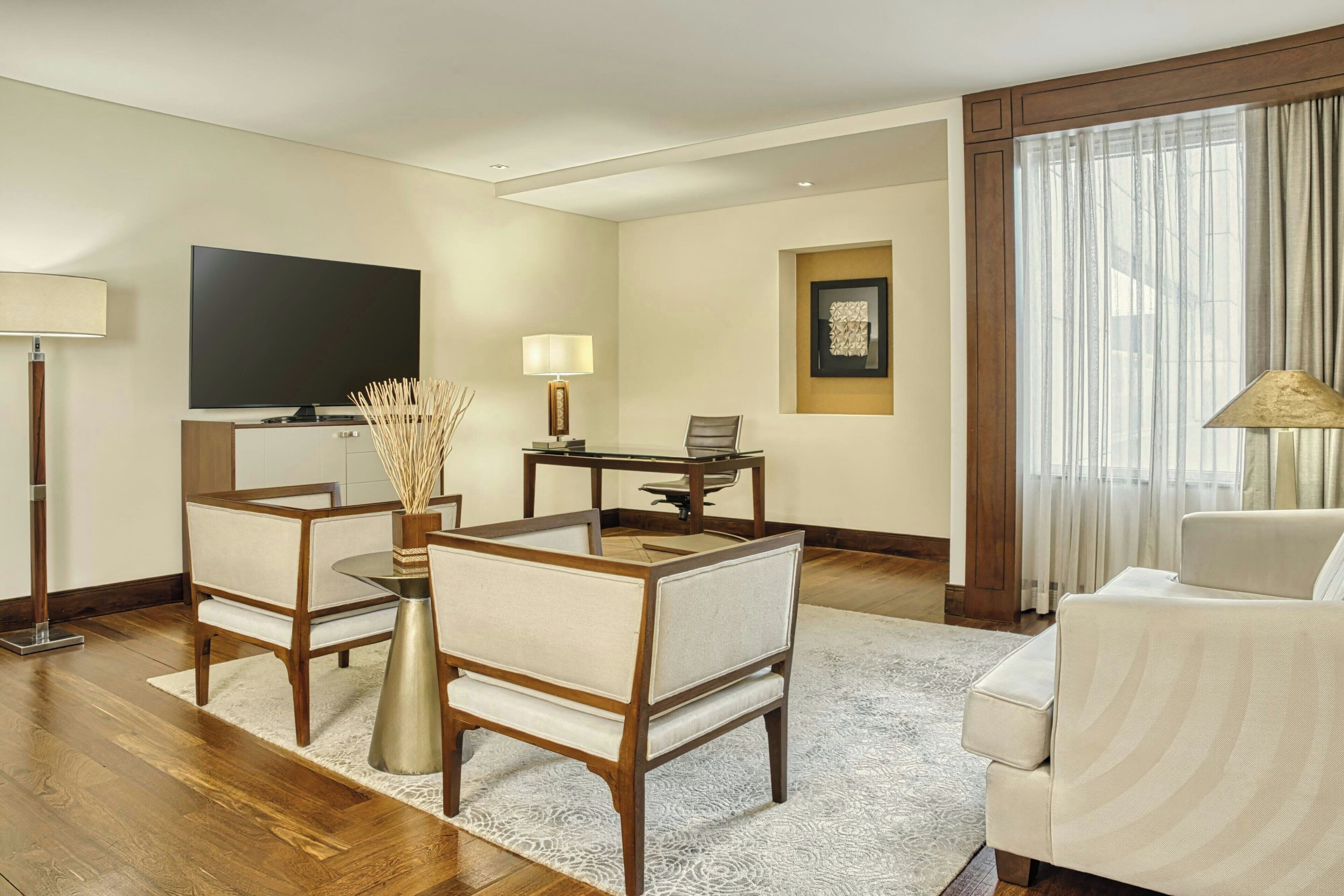 A hotel room with a flat-screen TV, a dining table with chairs, a sofa, a lamp, and a large window with curtains.