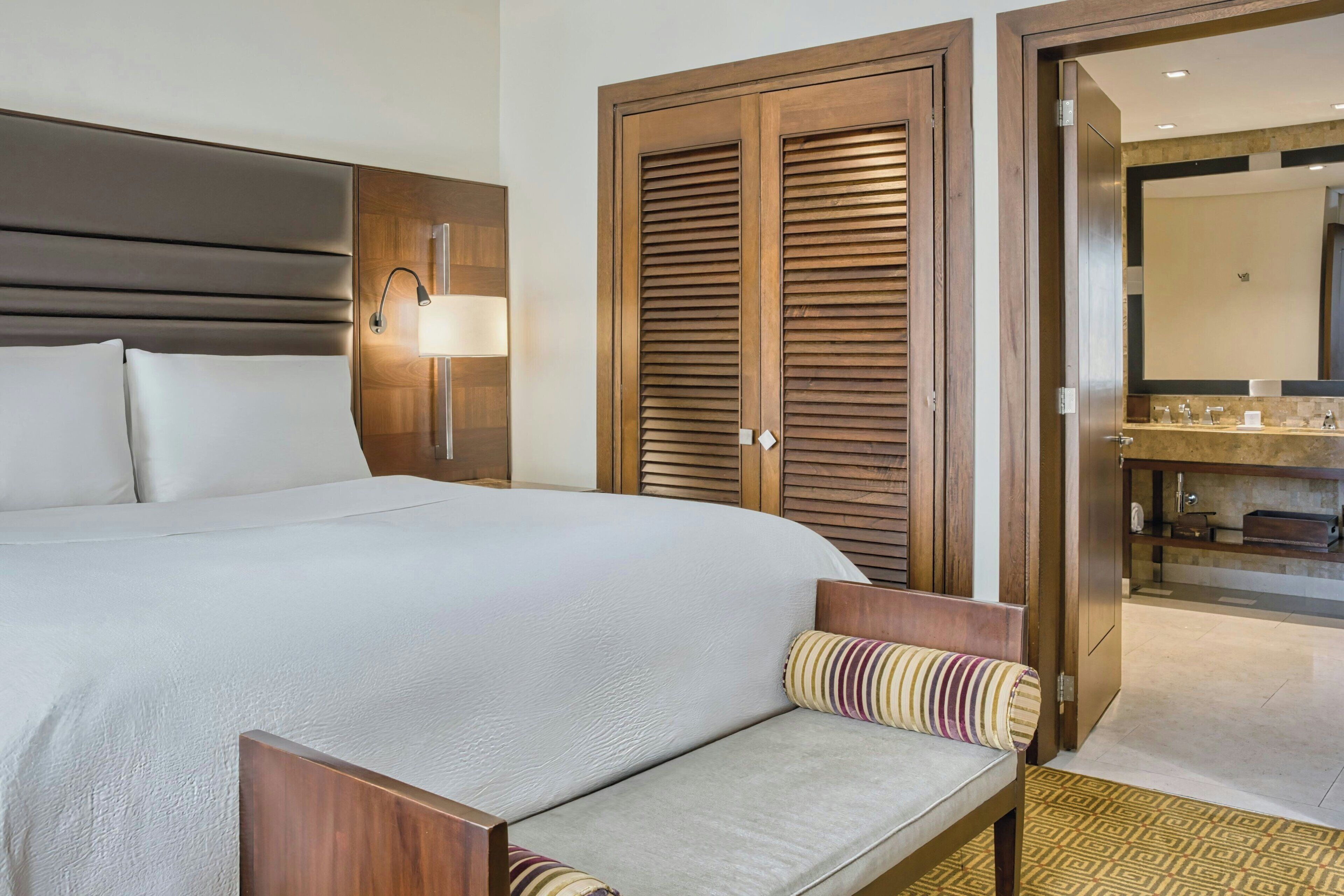 Ein Hotelzimmer mit einem großen Bett, einem Nachttisch mit Lampe, einem Holzschrank und einem durch eine offene Tür einsehbarer Badezimmer.