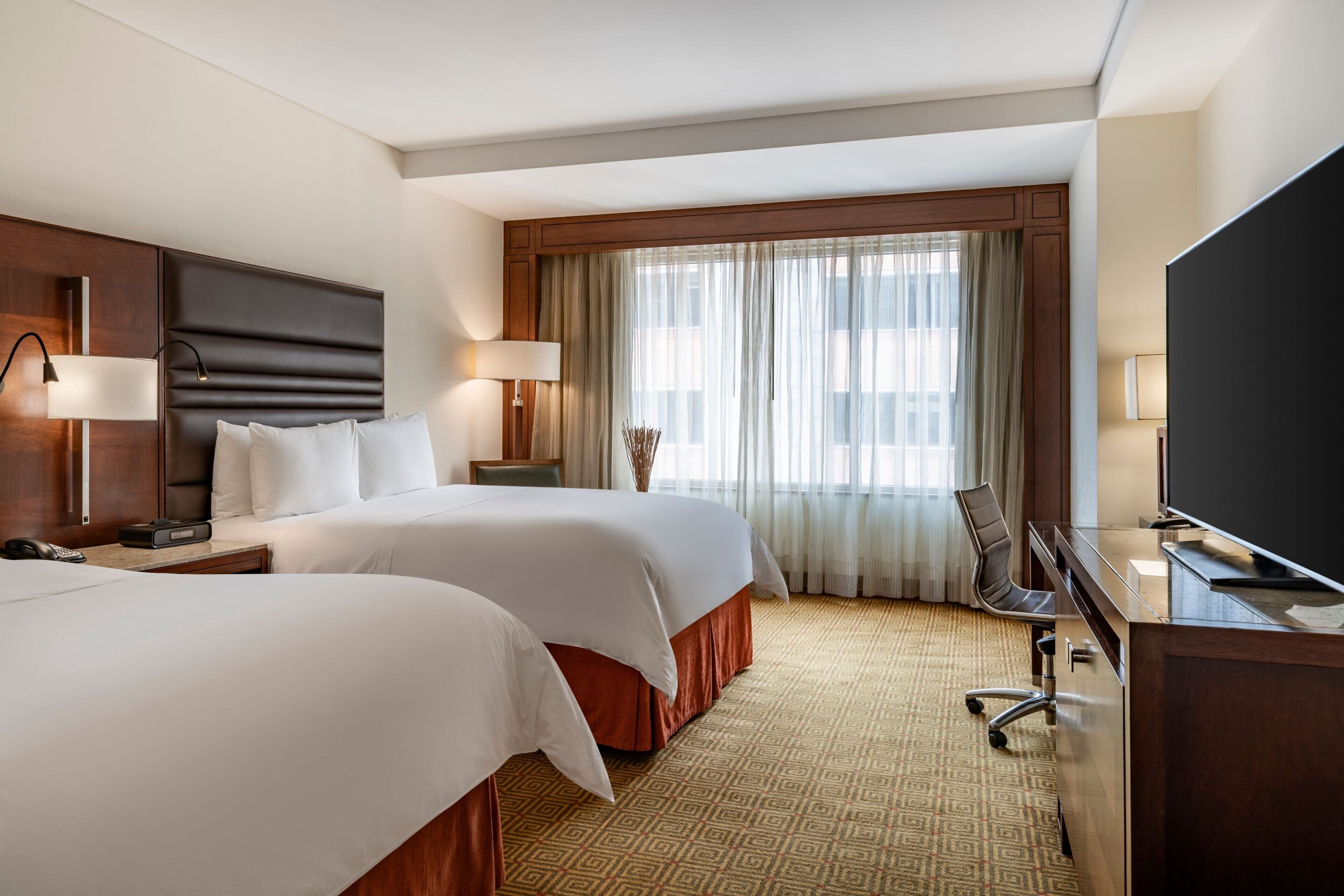 Quarto de hotel com duas camas, uma escrivaninha com televisão e uma janela grande com cortinas.