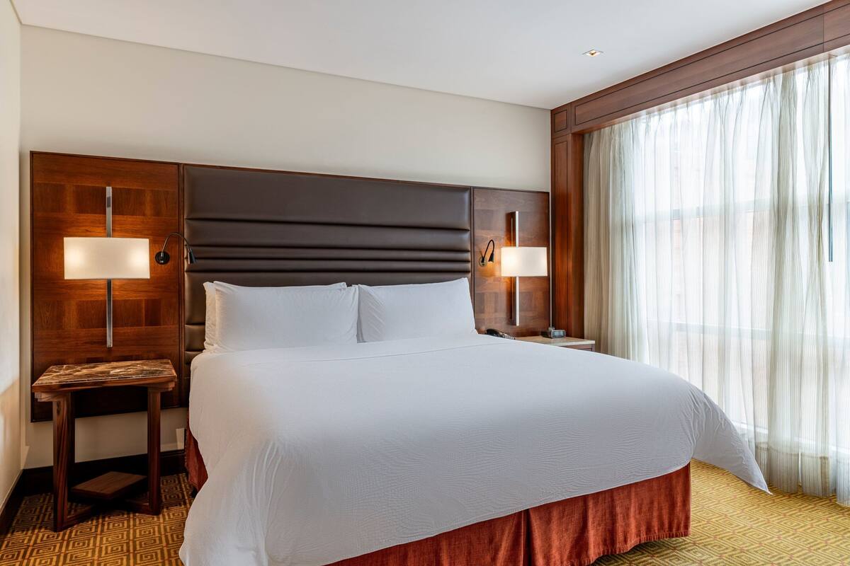 A hotel room with a large bed, two bedside tables with lamps, a wooden headboard, and a large window with sheer curtains.