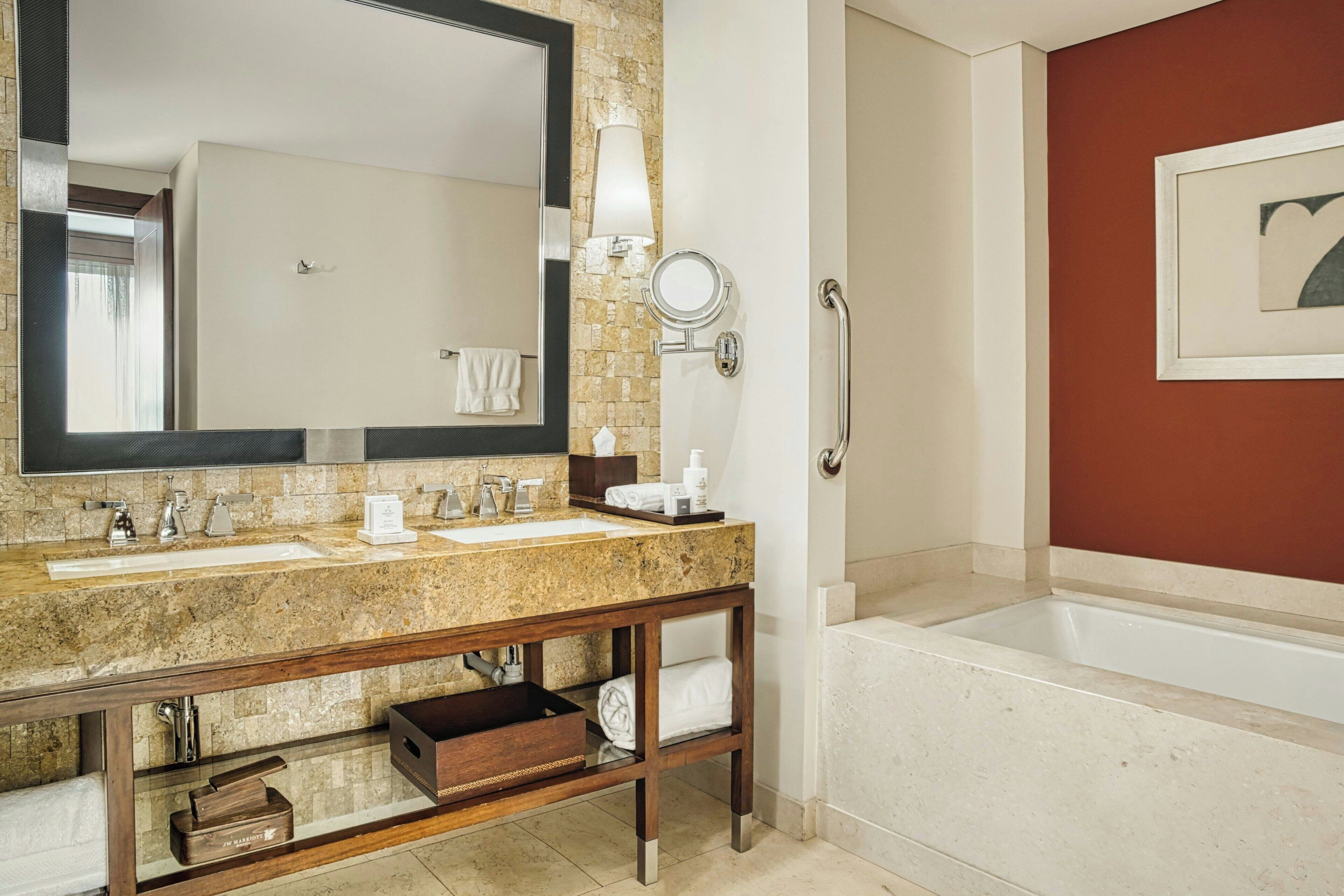 A bathroom with a double sink, a large mirror, a vanity with a wooden base, a towel rack, and a bathtub.