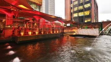 A waterfront restaurant with outdoor seating, a water feature, and modern buildings in the background.