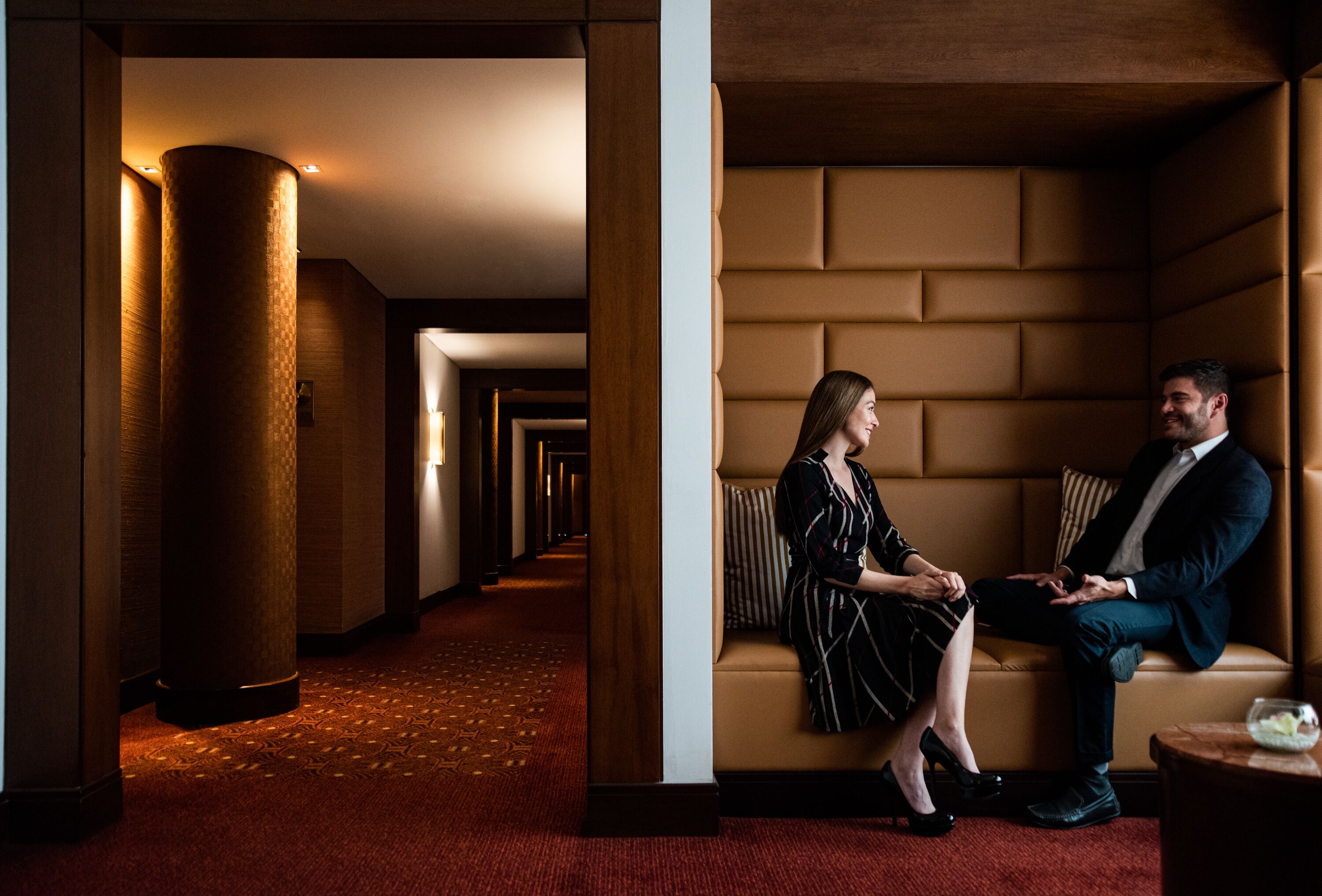 Un vestíbulo de hotel con un largo pasillo, una zona de estar con un sofá y una mesa redonda, y un pilar decorativo.
