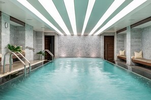 An indoor swimming pool with a stone wall backdrop, metal railings, and seating areas with cushions.