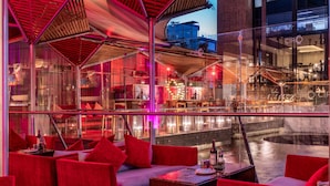 Un salón interior moderno con asientos rojos, barandilla de cristal y vista a un edificio a través de una fuente de agua.