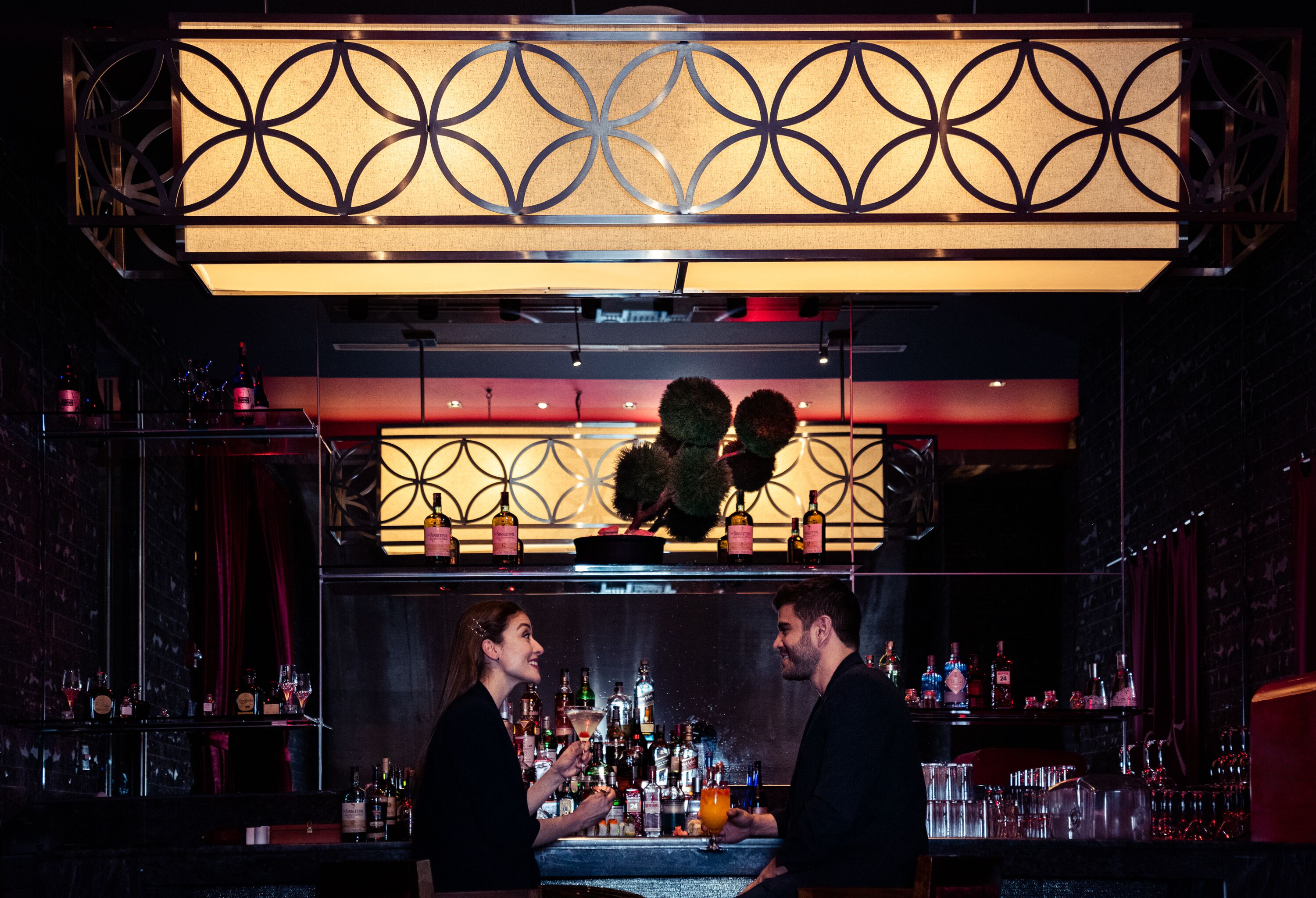 A bar area with a decorative overhead light fixture, shelves with bottles, and a counter where two people are standing.
