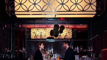 A bar area with a decorative overhead light fixture, shelves with bottles, and a counter where two people are standing.