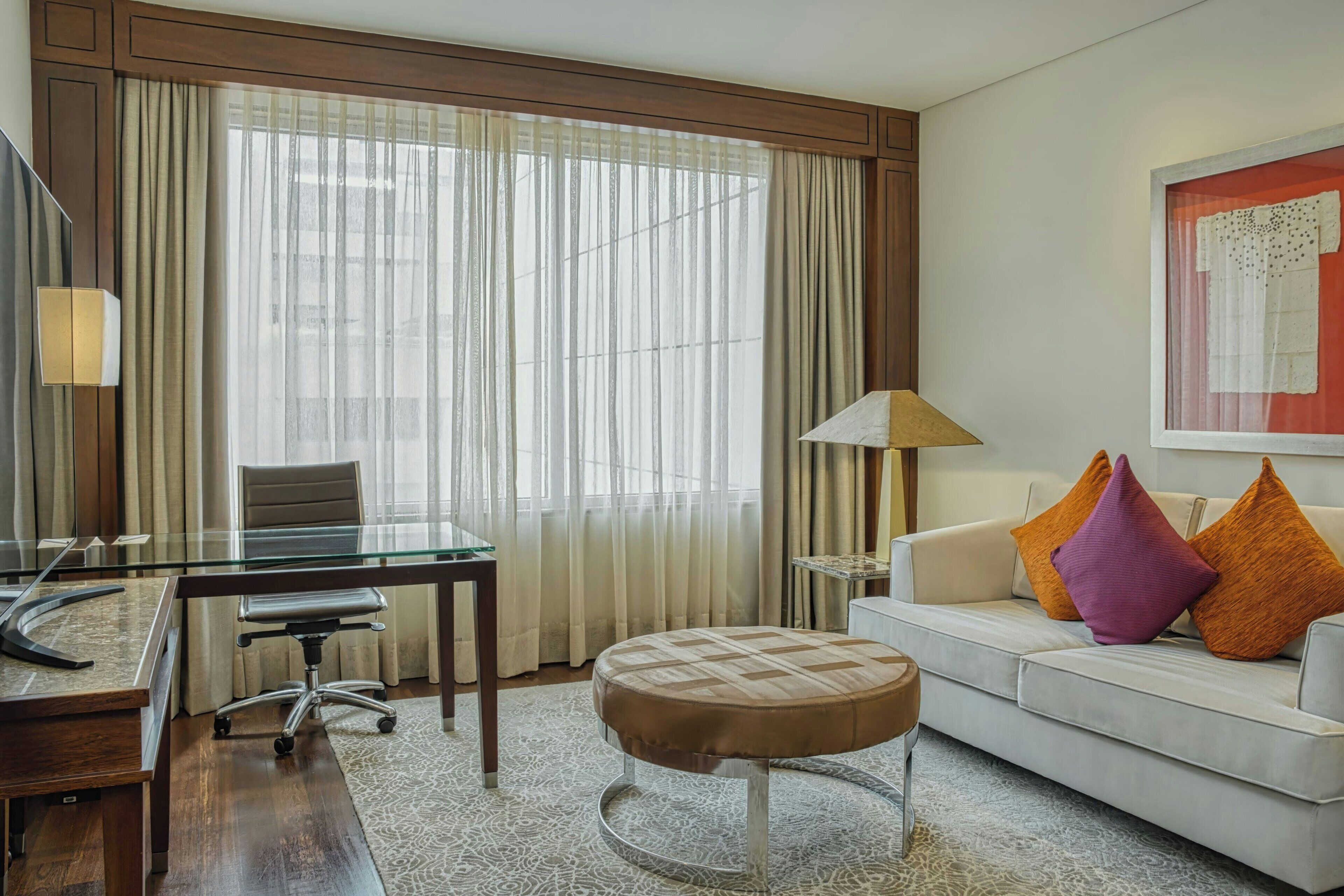 A hotel room with a glass table, a sofa with colorful cushions, a round ottoman, a lamp, and a large window with curtains.
