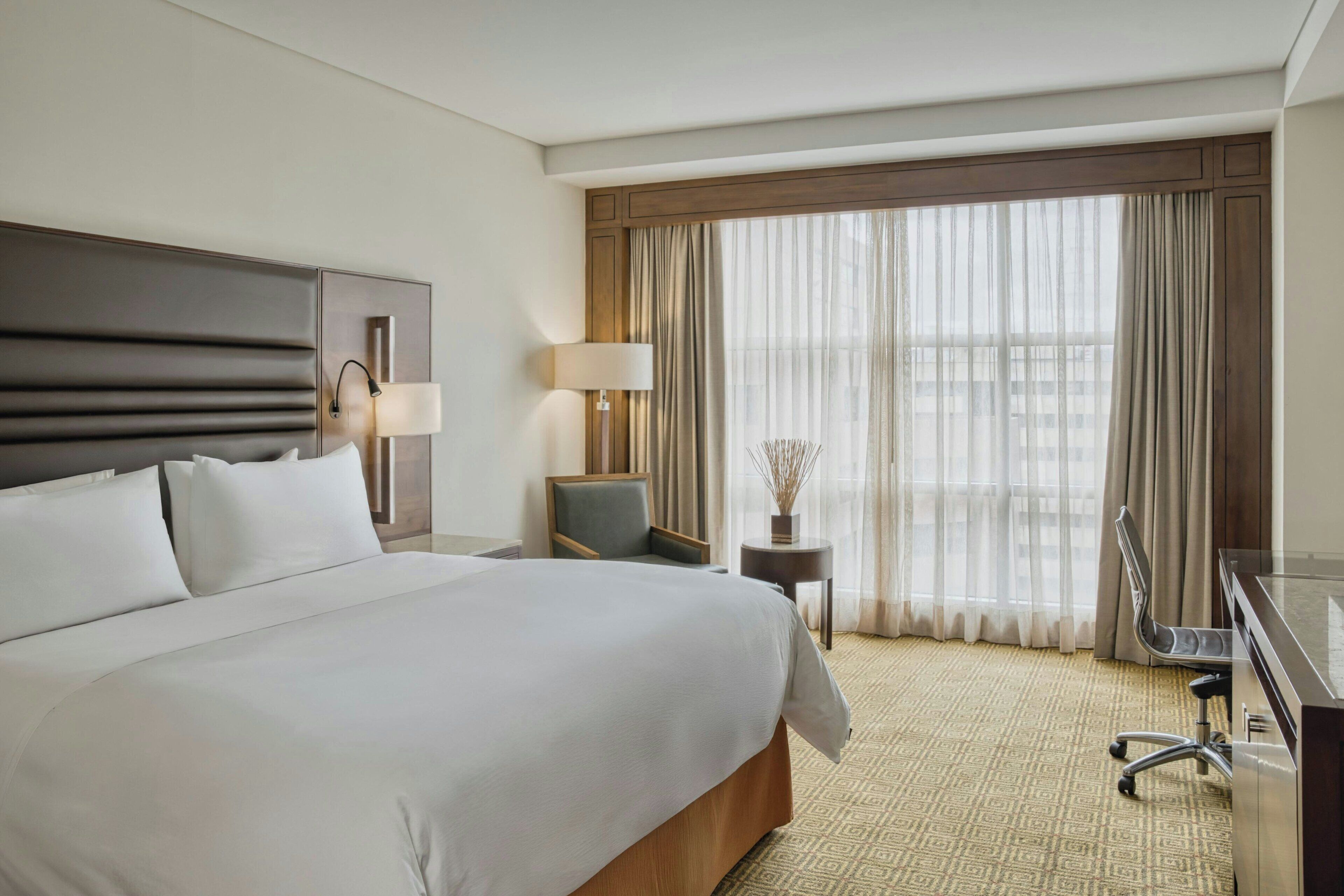 Ein Hotelzimmer mit einem großen Bett, einem Schreibtisch, einem Stuhl, einer Lampe und Blick aus dem Fenster.