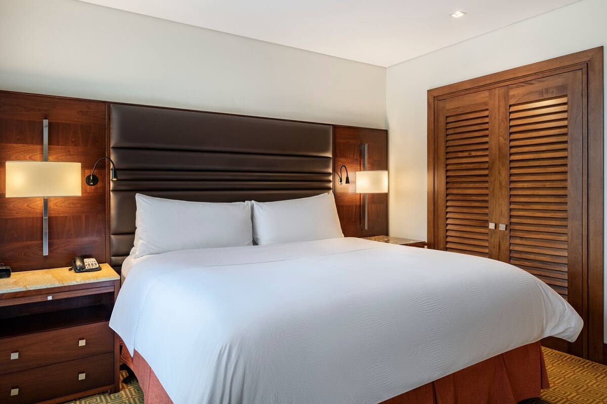 A hotel room with a large bed, wooden headboard, two bedside lamps, a wooden wardrobe, and a small desk with a telephone.