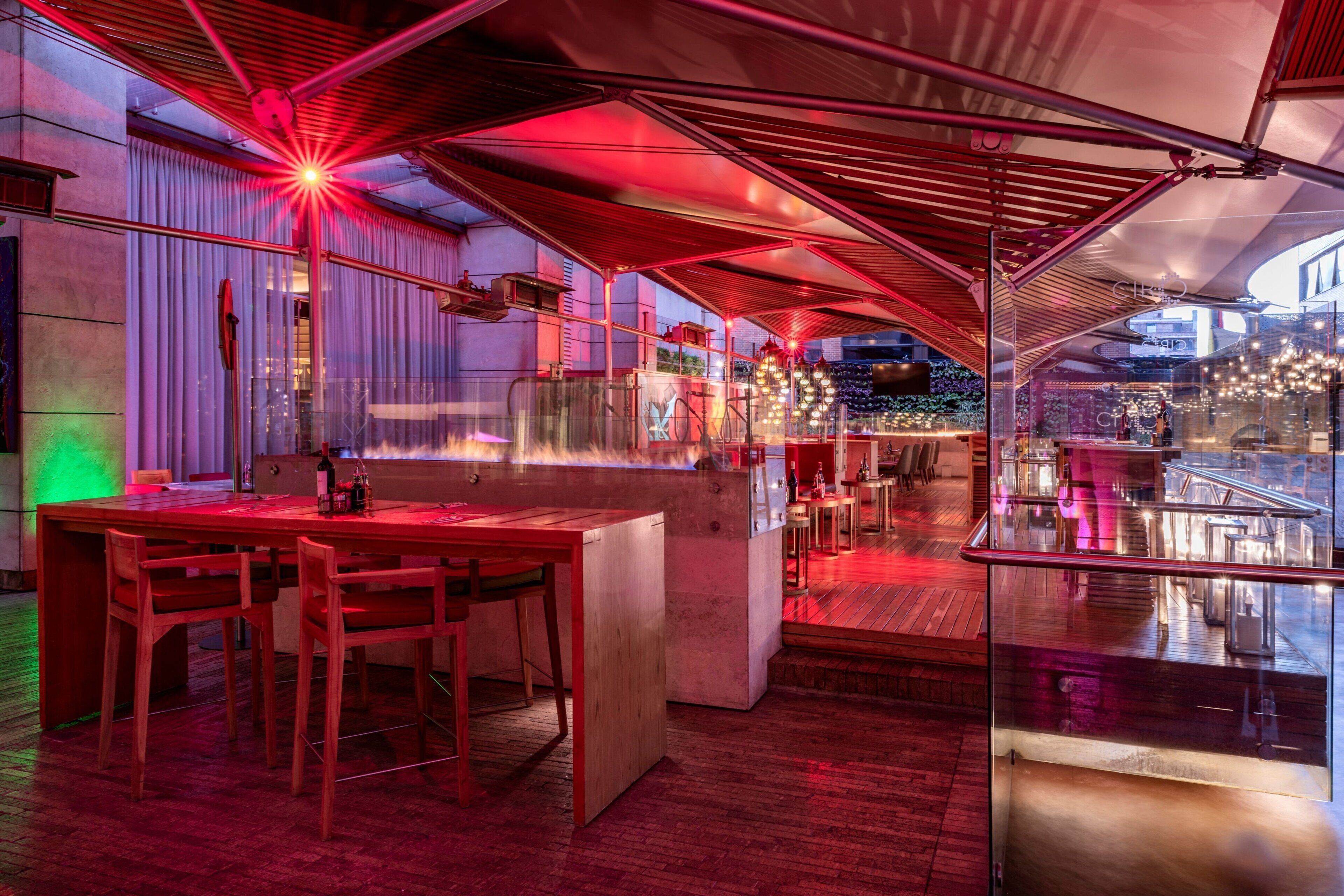 Zona de bar moderna con cerramiento de vidrio, sillas de madera y techo iluminado con luz roja.