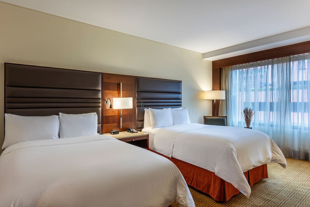 A hotel room with two beds, a wooden headboard, a nightstand with a lamp, and a window with sheer curtains.
