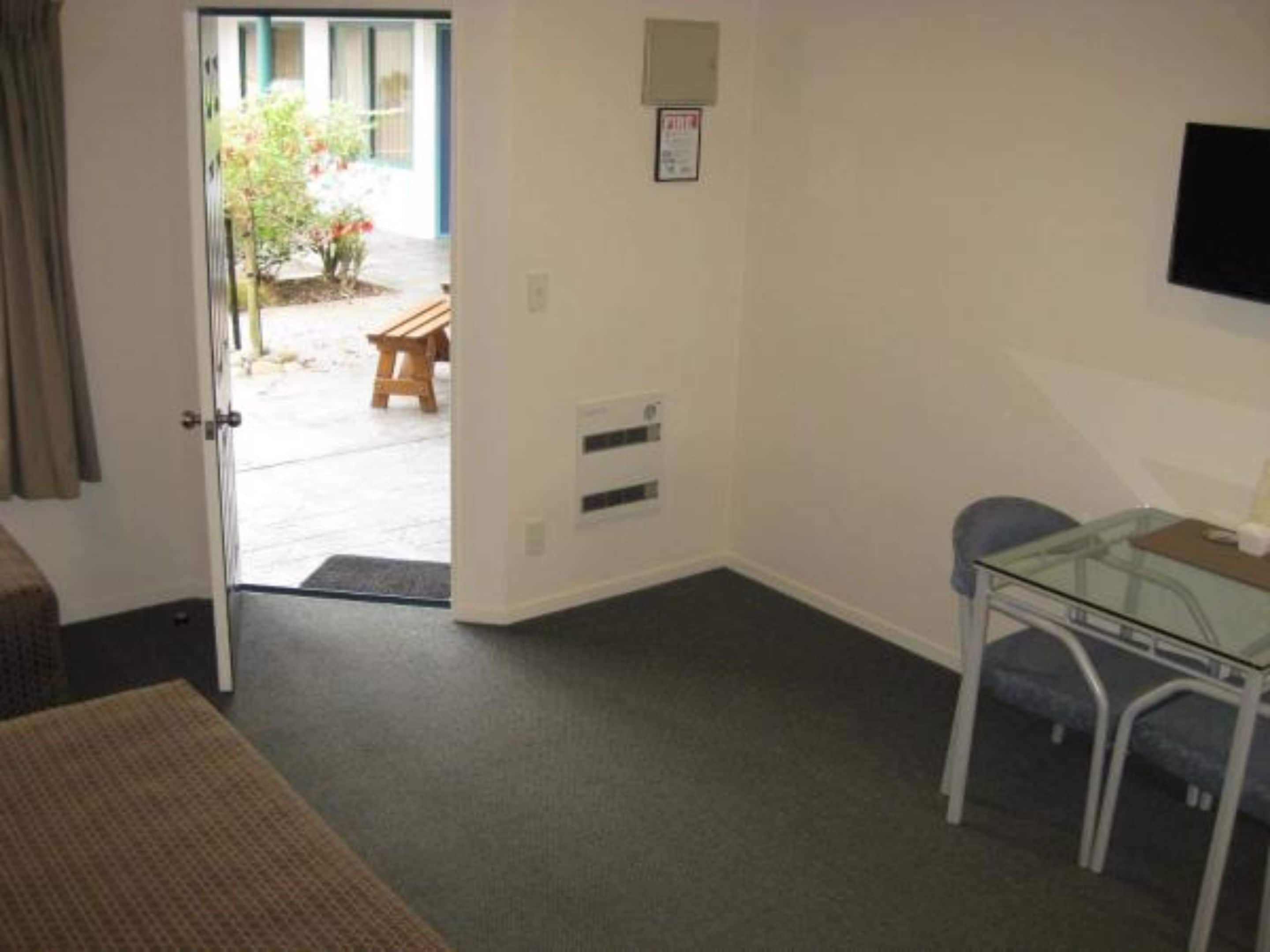 A room with a bed, a glass table, and a chair. There is a door leading to an outdoor area with a bench and plants.